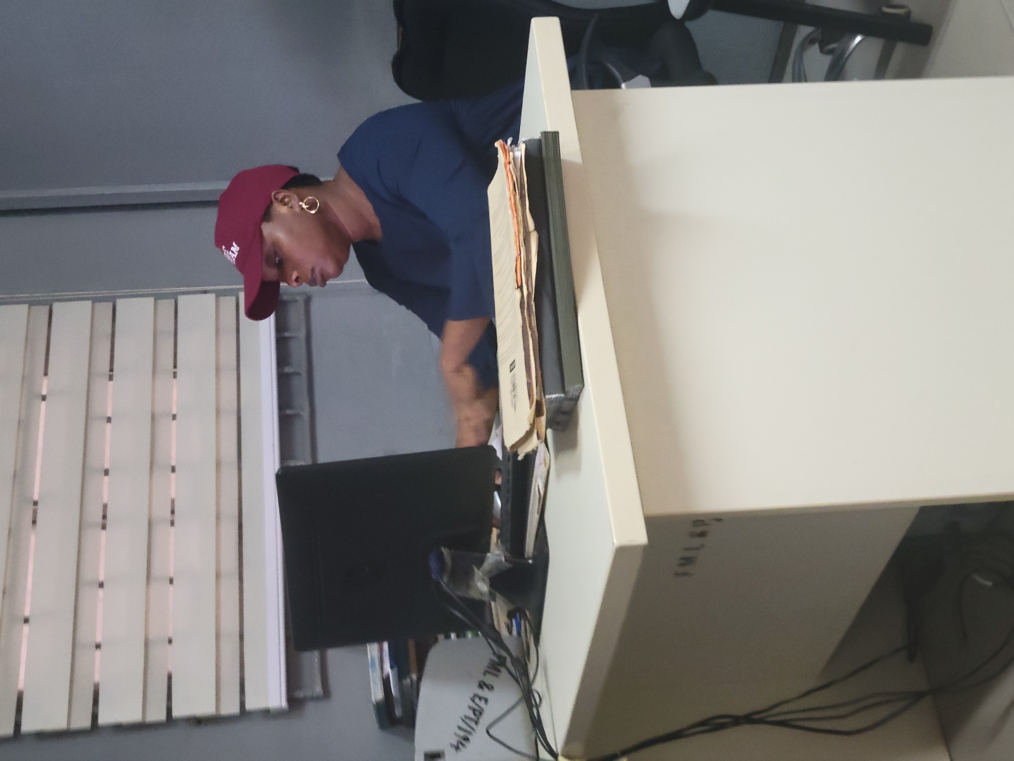 The image depicts a person in an office or workspace, potentially located in Abuja, Nigeria. The central figure is a dark-skinned individual, likely a woman, wearing a dark blue t-shirt and a maroon baseball cap. She is adorned with large gold hoop earrings. The person is captured bending forward, looking down with concentration at a black computer monitor and the immediate desk area.
The setting appears to be a cubicle or partitioned workspace, defined by a light beige or off-white desk and partition walls. To the left, a set of vertical blinds covers what appears to be a window, indicating an indoor, possibly daytime, environment.
On the desk surface, near the monitor, a document holder filled with various papers is visible. Multiple cables are seen connected to the monitor and other unseen equipment. Text is discernible on various elements within the frame: "FM L&P?" and "FML&P" are written on the desk partition, while a label or tag below the monitor clearly shows "PML & E/PT/194". The activity suggests the person is engaged in work or examining something at the computer workstation. No specific emotions are clearly evident beyond a sense of focus.