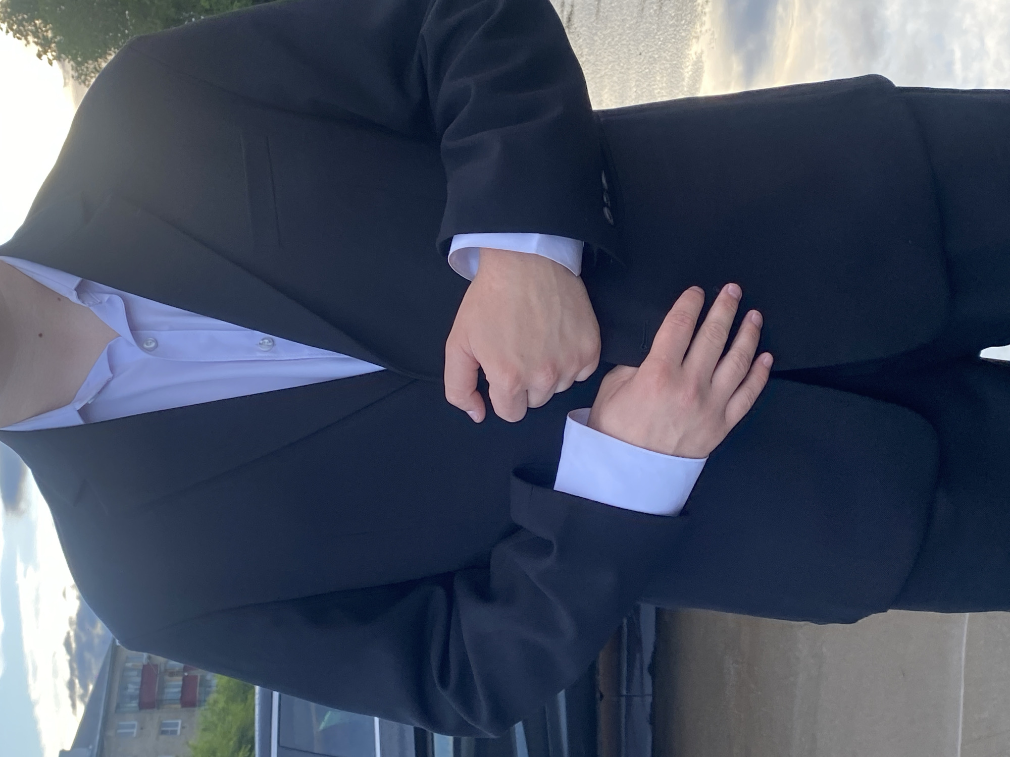 A person wearing a black suit jacket and a light blue dress shirt is the subject of this photograph. The jacket is buttoned, and the shirt collar is visible. The person's hands are clasped in front of them, with the right hand resting on top of the left. The image is a close-up shot, focusing on the torso and arms of the person.
The background suggests an outdoor setting with a cloudy sky and some greenery visible on the left side of the frame. In the distance, a building with red accents can be seen, hinting at an urban or suburban environment. The lighting indicates it is daytime, possibly late afternoon or early morning due to the soft light and the presence of clouds. The overall mood appears neutral, with no discernible emotions conveyed by the subject. There is no visible text in the image. The setting context of Uchaly, Russia, is provided.