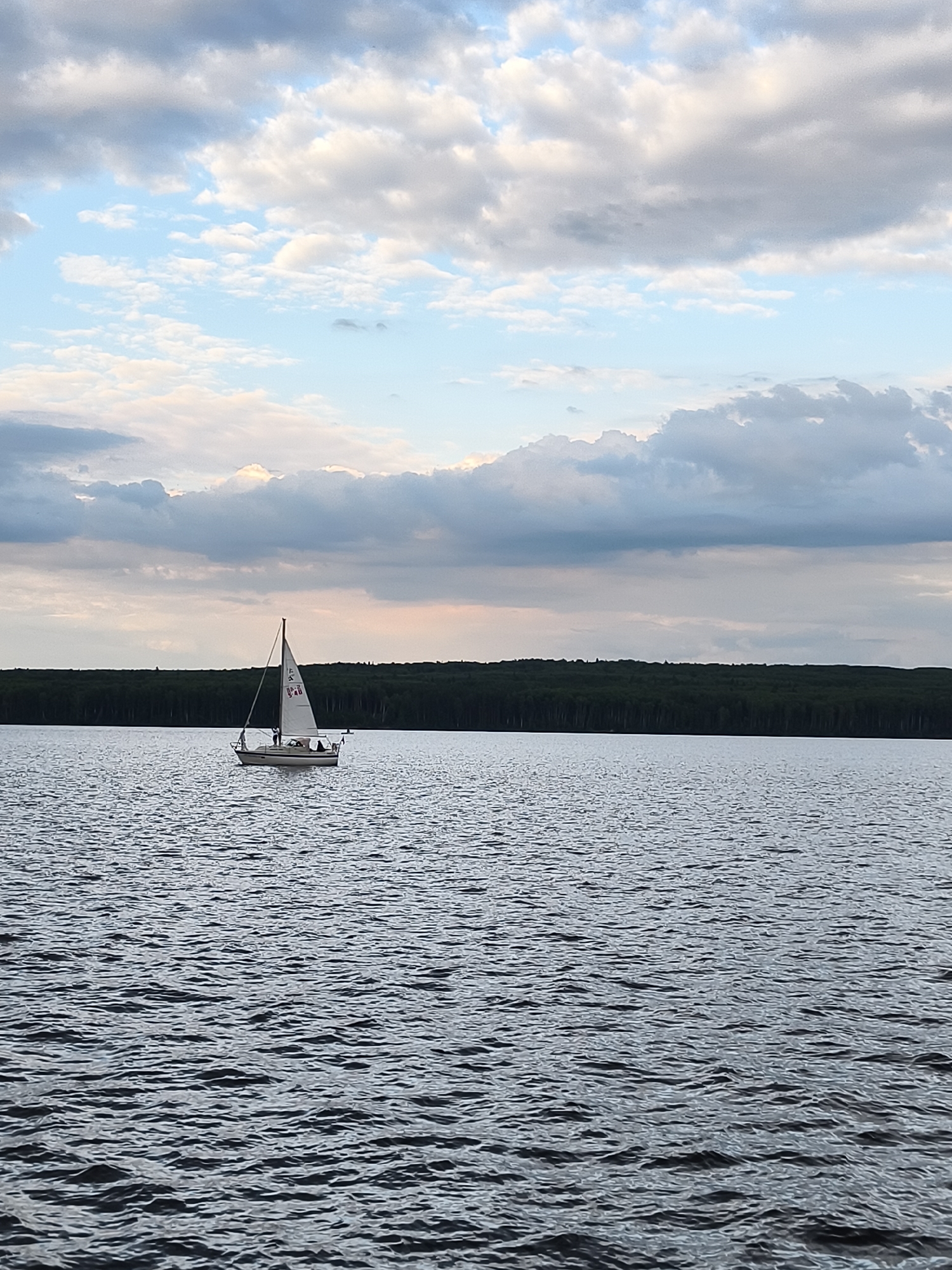 This media file captures a serene scene on a body of water, likely a lake or calm sea, with a sailboat as the central object. The sailboat, white-sailed and relatively small, is positioned in the middle of the frame, indicating it is underway. A few indistinct figures can be seen on its deck, suggesting human activity.
The background features a dense, dark green treeline that stretches horizontally across the image, suggesting a natural, forested landscape. Above the treeline, the sky is a dynamic mix of blues and grays, with scattered clouds that appear to be moving, possibly indicating a breezy or changeable weather condition. The time of day appears to be late afternoon or early evening, given the soft lighting and the hues in the sky. The water is choppy, with numerous small waves and ripples reflecting the cloudy sky, adding texture to the scene.
There are no explicit location cues within the image itself, but the provided context places it in Chaikovsky, Russia. No visible text is present on the sailboat or in the surrounding environment within the frame. The overall mood of the image is peaceful and contemplative, highlighting the solitary beauty of sailing and nature.