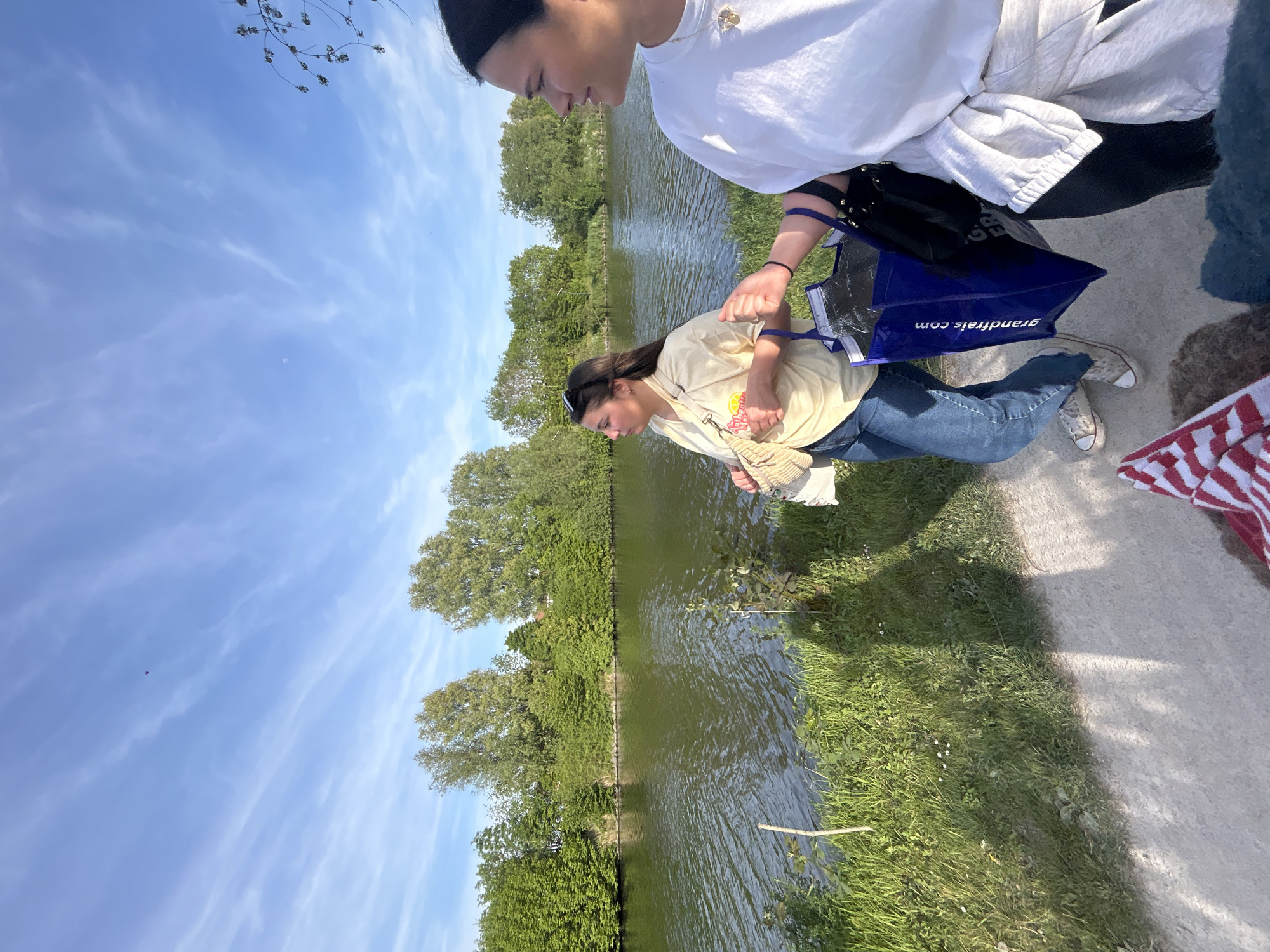 The image depicts two people standing on a path next to a canal or waterway in what appears to be a park. The location is Marquette-lez-Lille, France.
One person, wearing a white top and dark bottoms, is partially visible at the top of the frame. The second person, below them, is wearing a light-colored t-shirt and blue jeans, and holds a blue shopping bag with the name "grandfrais.com" printed on it. They also carry a beige handbag. Both individuals are looking downwards, seemingly at the water.
The waterway is lined with green vegetation, with trees along the far bank. The sky is blue with wispy clouds. Part of a red and white striped object is visible on the right side of the path.