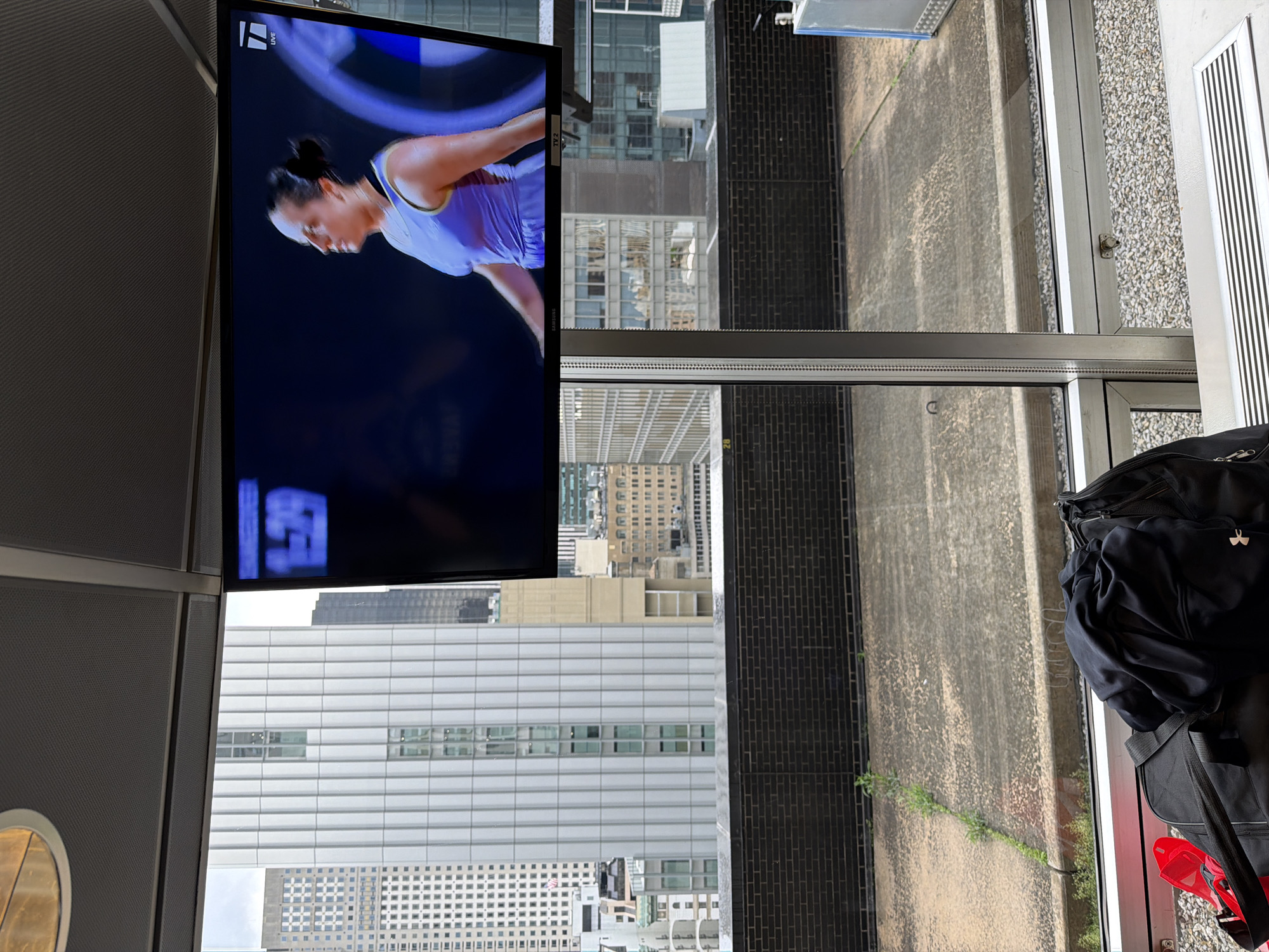 View from a high-rise building in New York City, United States. In the foreground, a television screen displays a female athlete in action, with a clear cityscape visible through the window behind. Below, a mix of older and more modern high-rise buildings forms the urban landscape. Next to the window, personal items, including clothing and a duffel bag, are present, indicating the scene is indoors, possibly an apartment or hotel room.