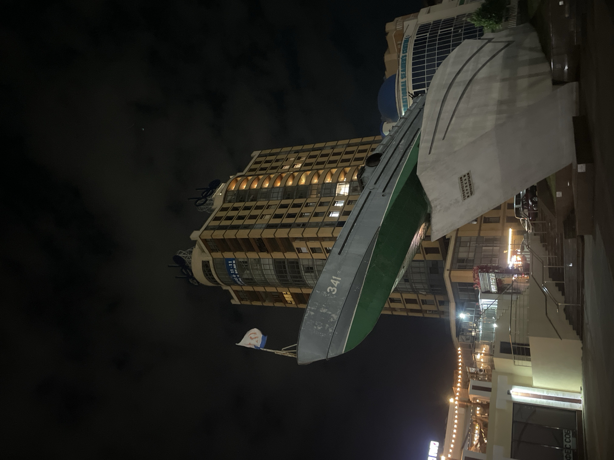 This nighttime image captured in Varnavinskoye, Russia, features a striking monument of a submarine, likely a memorial or landmark, prominently displayed in front of a modern, illuminated high-rise building. The submarine, painted gray with a green hull, is mounted on a concrete base that is designed to resemble the bow of a ship. A flag, predominantly white with blue and red elements and a circular emblem, flies from the submarine's periscope. The number "341" is visible on the side of the submarine.
The tall building behind the submarine has a multi-story facade with numerous windows, some of which are lit, suggesting occupancy. The building's architecture is modern, with a curved top section and several antenna-like structures. To the right of the main building, another structure with a blue dome is partially visible. In the foreground, a paved area with stairs and railings suggests a public access point. Scattered lighting fixtures, including strings of lights on a lower building to the left, illuminate the scene. The sky is dark and appears to be cloudy, with faint hints of stars or distant lights. No people are clearly visible in the image, and there are a few parked vehicles in the background. The overall scene suggests a public space with a focus on maritime history or commemoration.