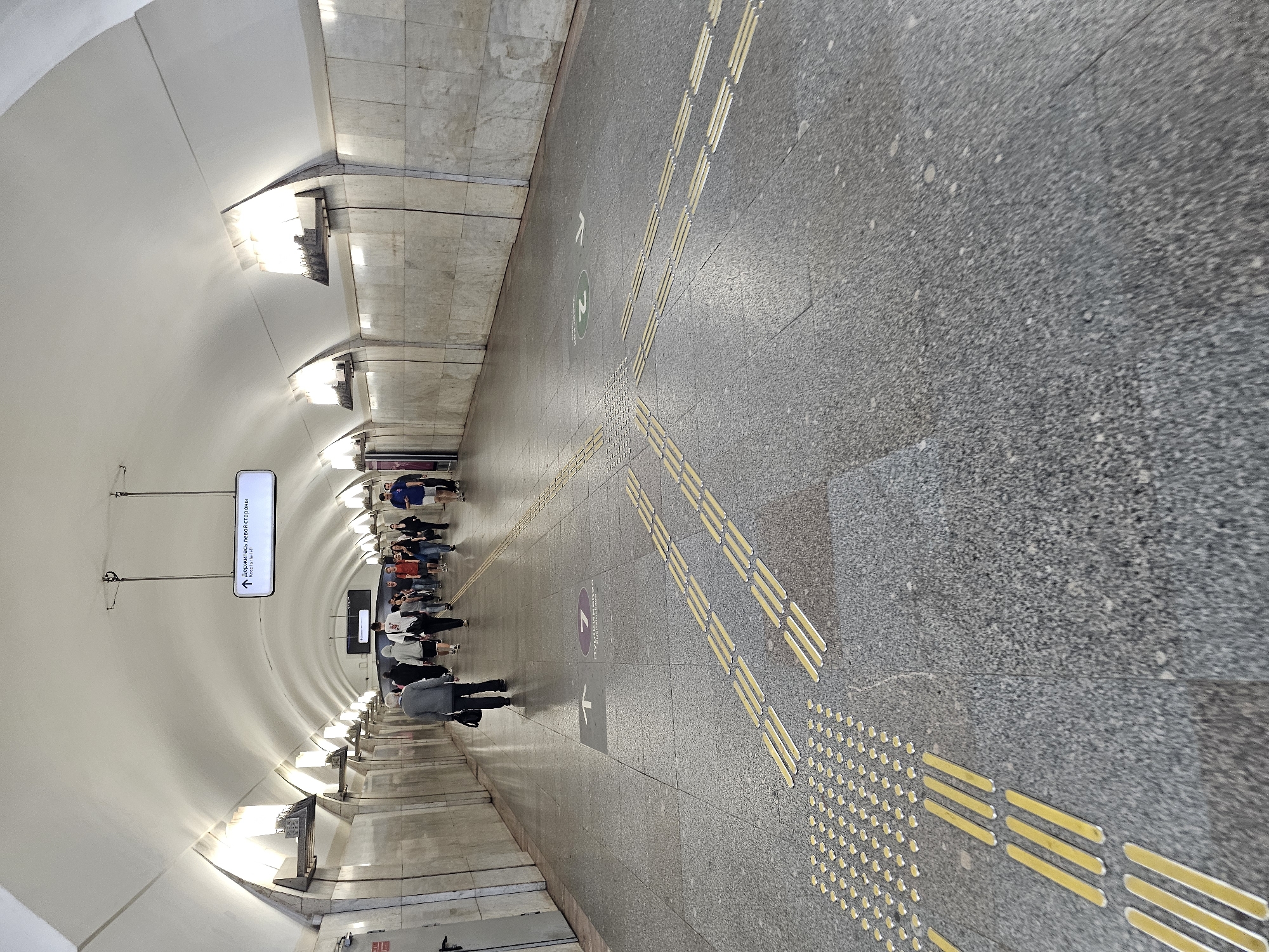The image depicts a long, vaulted pedestrian tunnel, likely within a Moscow metro station, given the architectural style, signage, and the presence of tactile paving for the visually impaired. Several people are walking in both directions along the tunnel. The walls are lined with light-colored marble tiles, and the ceiling features an arched structure with integrated lighting fixtures. Yellow tactile paving strips and discs are visible on the floor, guiding pedestrians. Signage is present, including one sign with an arrow pointing forward and Cyrillic text indicating "Держитесь левой стороны" (Keep to the left side), and another sign with a green circle and the number "2" with an arrow. The floor is made of grey speckled granite tiles. The overall atmosphere is that of a busy underground public space.