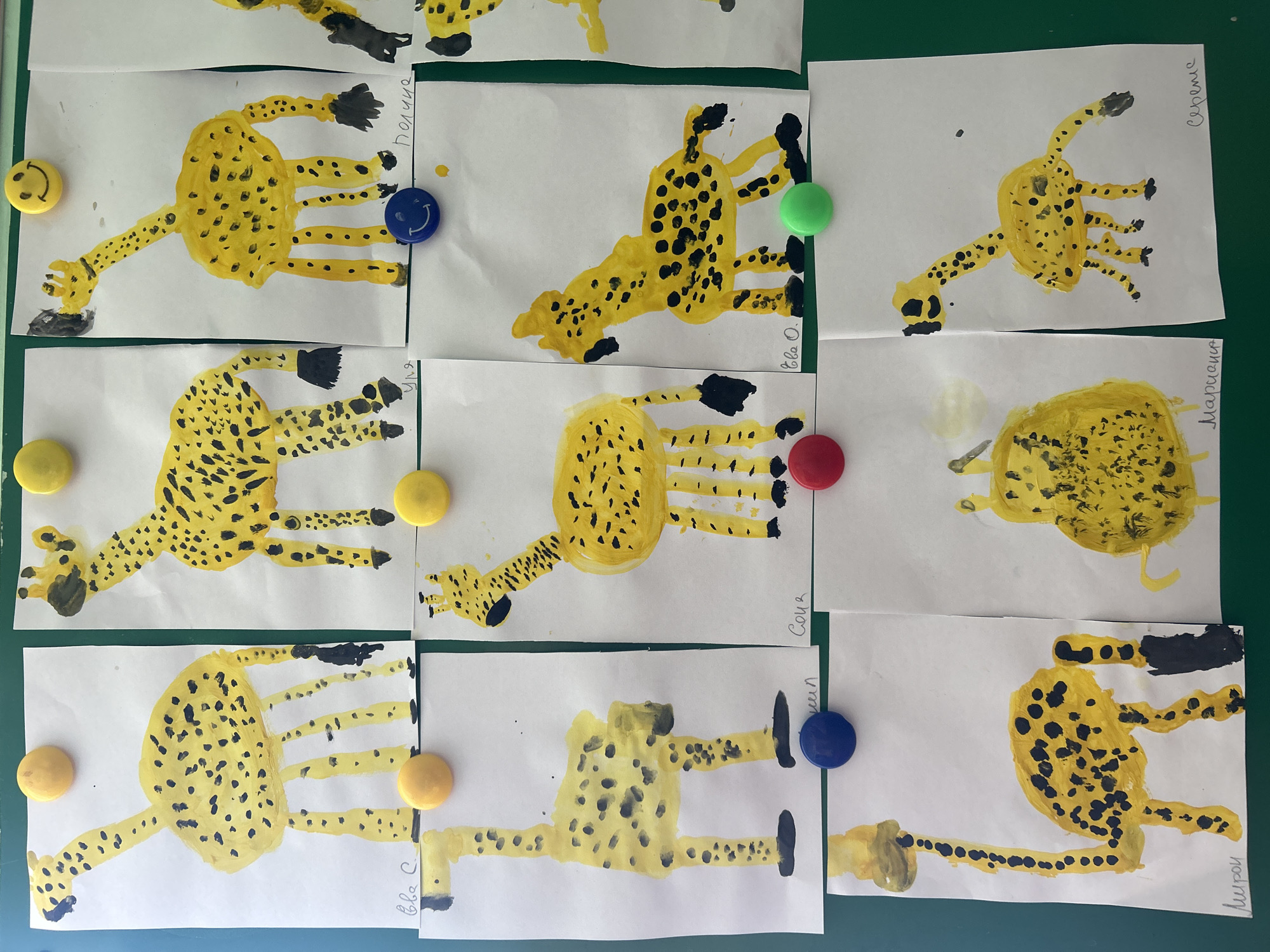 The image shows a collection of nine children's drawings of giraffes, each attached to a green surface, possibly a bulletin board or wall. The drawings are rendered in yellow paint with black spots, depicting giraffes in various poses. Each drawing is affixed with a colorful magnetic pin: yellow, blue, green, or red. The names "Полина" (Polina), "Ева О.", "Сергеие" (Sergeie), "Уля" (Ulya), "Соня" (Sonya), "Марианна" (Marianna), "Ева С." (Eva S.), "Семен" (Semyon), and "Мирон" (Miron) are handwritten next to or beneath the drawings, presumably identifying the artists. The scene appears to be in an indoor environment, likely a classroom or daycare in Chaikovsky, Russia.