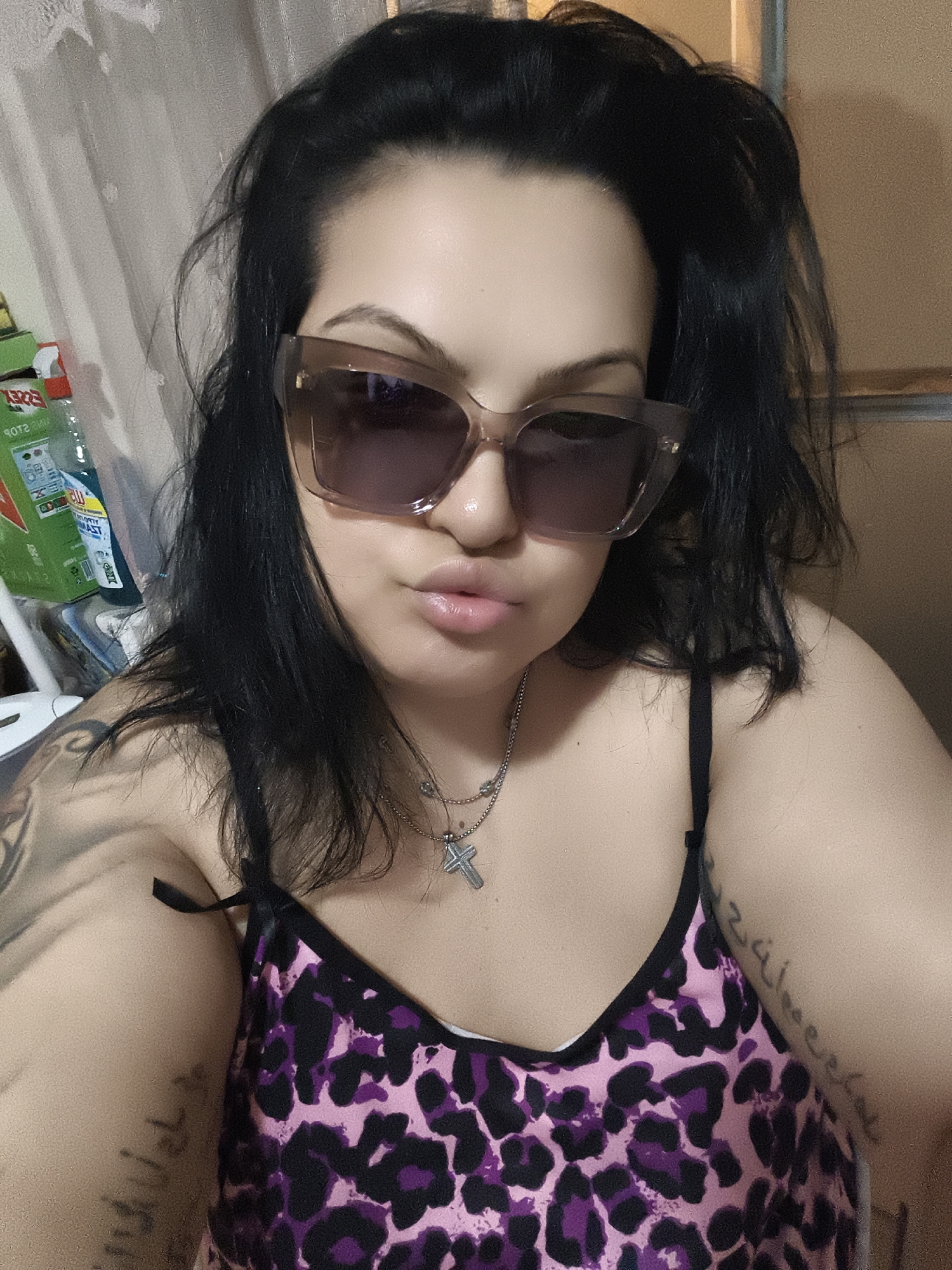 A woman with dark hair and sunglasses is taking a selfie in what appears to be a domestic setting, possibly her home. She is wearing a camisole with a pink and black leopard print. A silver necklace with a cross pendant is visible. There are tattoos on her arms and shoulder. In the background, there are some cleaning supplies and a curtain. The lighting is artificial, creating some shadows and highlights. The image does not provide any information to place it in Komotini, Greece.