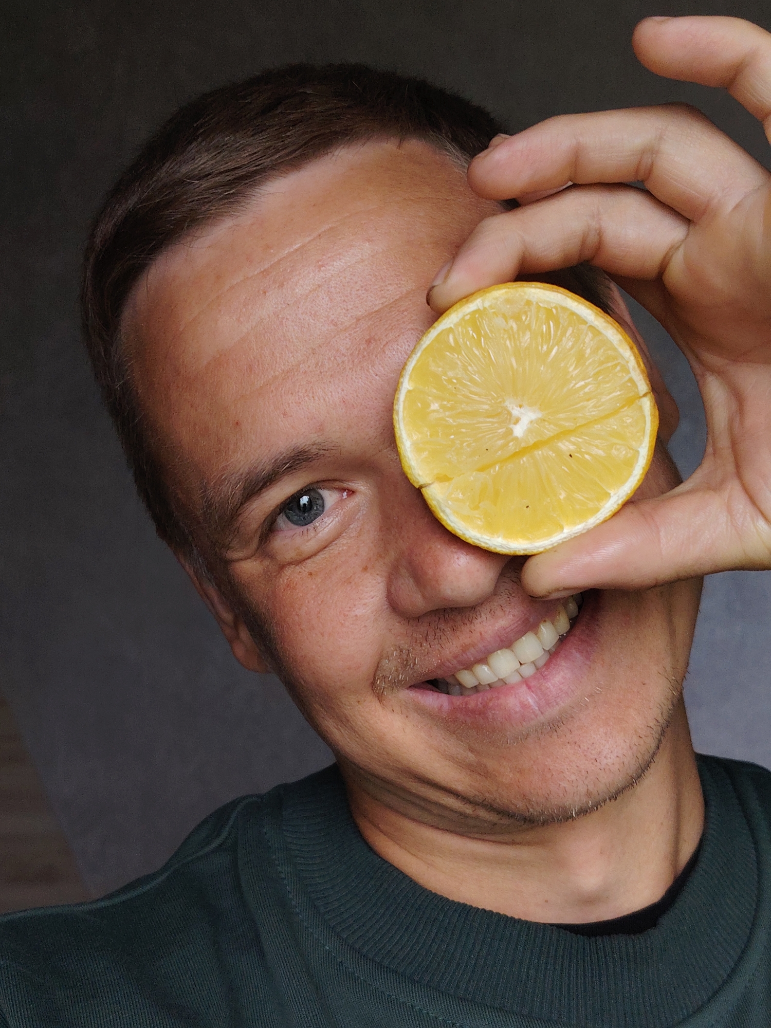 A close-up, eye-level shot features a smiling man holding a half-sliced yellow lemon over his right eye, partially obscuring his face. His left eye, which is blue, is clearly visible, conveying a playful expression. He has short, dark brown hair and light stubble on his chin and upper lip. He is wearing a dark green crew-neck sweater or shirt. The background is a plain, dark gray surface, suggesting an indoor setting. Natural light illuminates his face from the front-left, creating subtle shadows under his nose and on his neck. The man's mouth is open in a wide smile, revealing white teeth. His skin shows some fine lines on his forehead and around his visible eye. There is no visible text in the image. The overall mood is cheerful and light-hearted. The location context for this image is Chaikovsky, Russia.