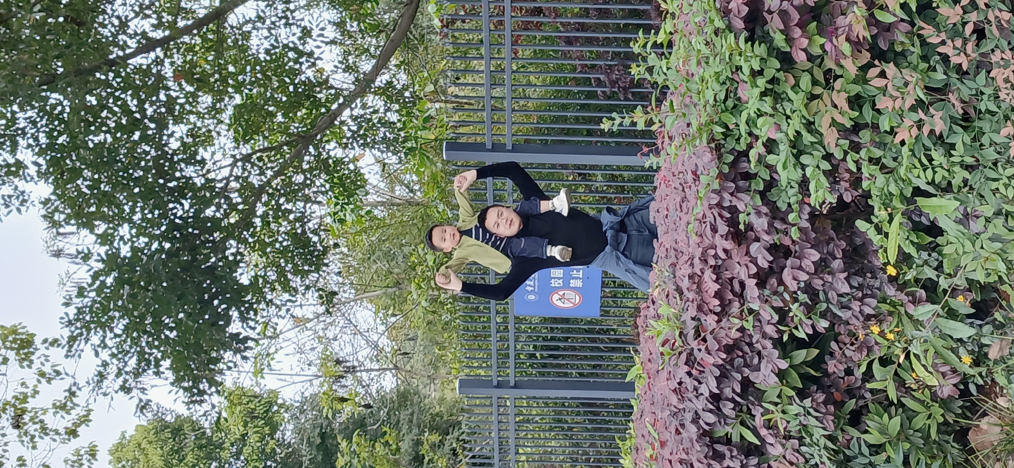 A man and a child are posing in front of a fence next to a bush in the Chongqing High-tech Zone, China. The man is wearing a black shirt and blue jeans, and the child is wearing a green shirt and white shoes. They are holding hands and smiling. A sign is visible on the fence. Trees surround the scene.