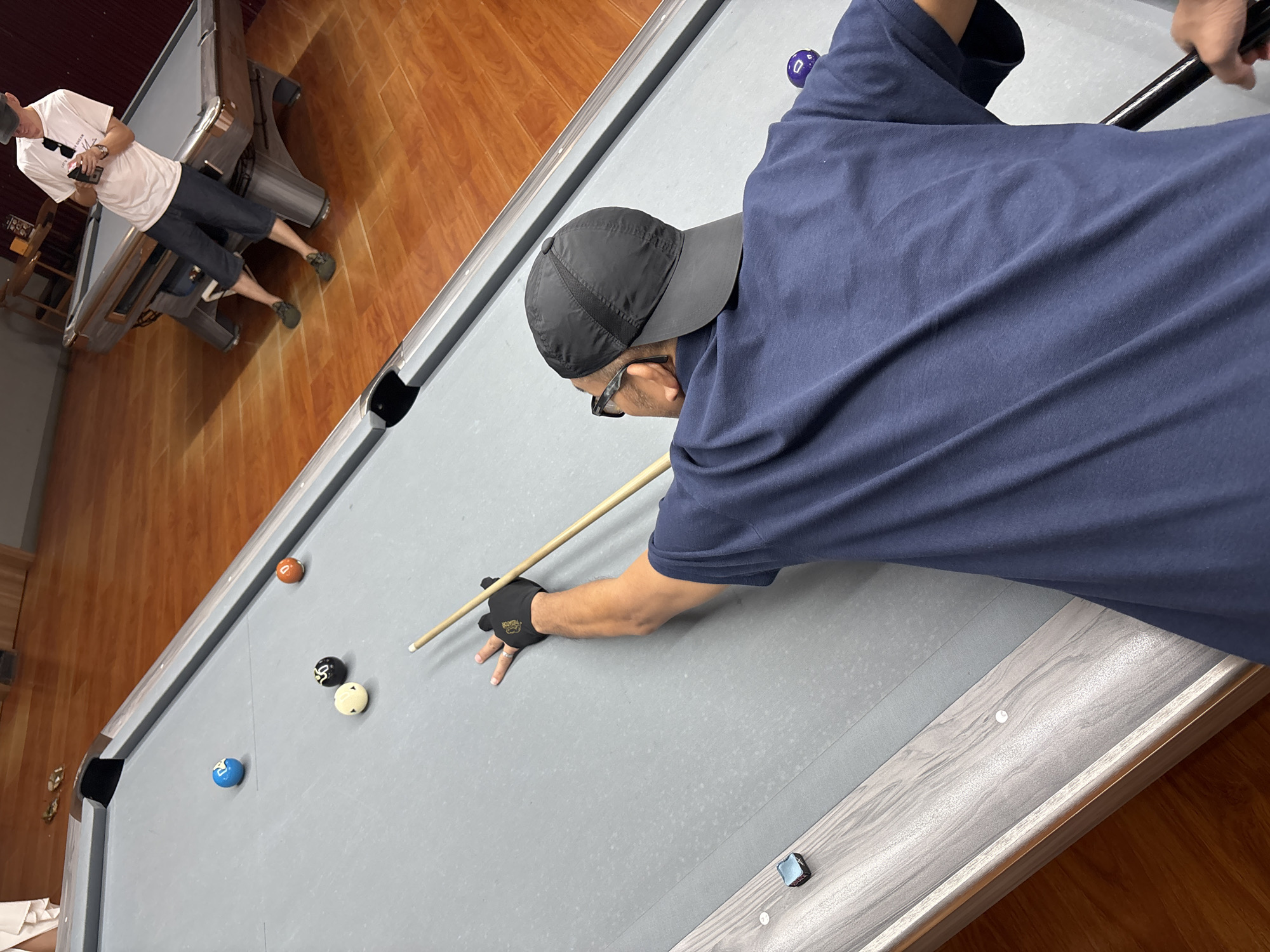 In this overhead shot, a person wearing a dark blue t-shirt, a black baseball cap, and sunglasses is playing pool. They are positioned on the right side of the frame, leaning over the pool table, holding a cue stick with their right hand, which is covered by a black glove. Their left hand rests on the table for support. Several pool balls are visible on the grey felt surface of the table: a purple ball numbered '0', an orange ball, a black and white ball, a white ball, and a blue ball. Another pool table is partially visible on the left side of the image. The setting appears to be an indoor pool hall with wooden flooring and walls. The lighting suggests it could be daytime, possibly indoors with artificial lighting. The scene captures a moment of focused concentration as the player prepares to strike the cue ball.