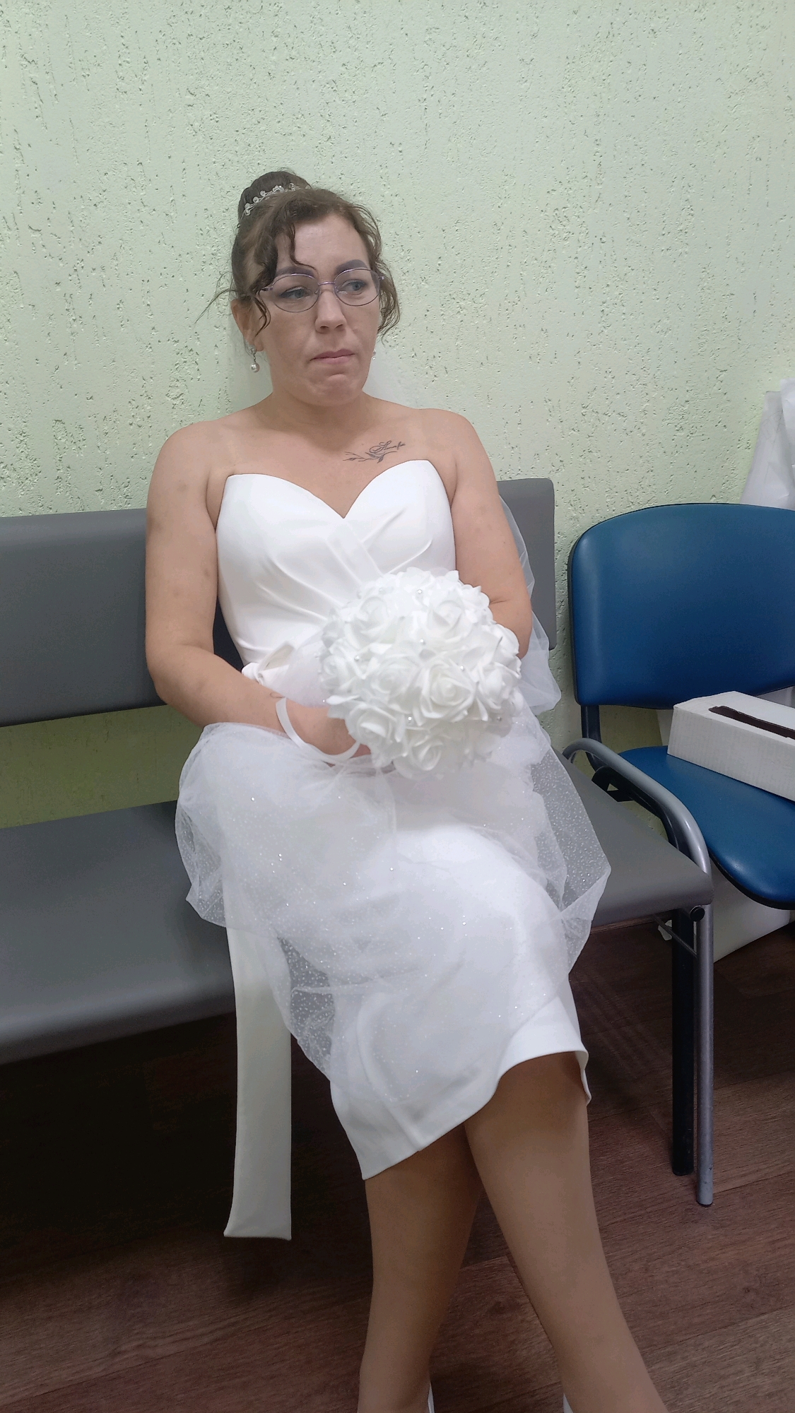A woman wearing a white strapless wedding dress is seated on a gray bench. She has her hair pulled up and is wearing glasses and earrings. She is holding a white bouquet of roses. The background is a textured light green wall. To the right of the bench is a blue chair with a white tissue box on it. The floor is made of dark wood planks. This scene is located in Pervozvanivka, Ukraine.