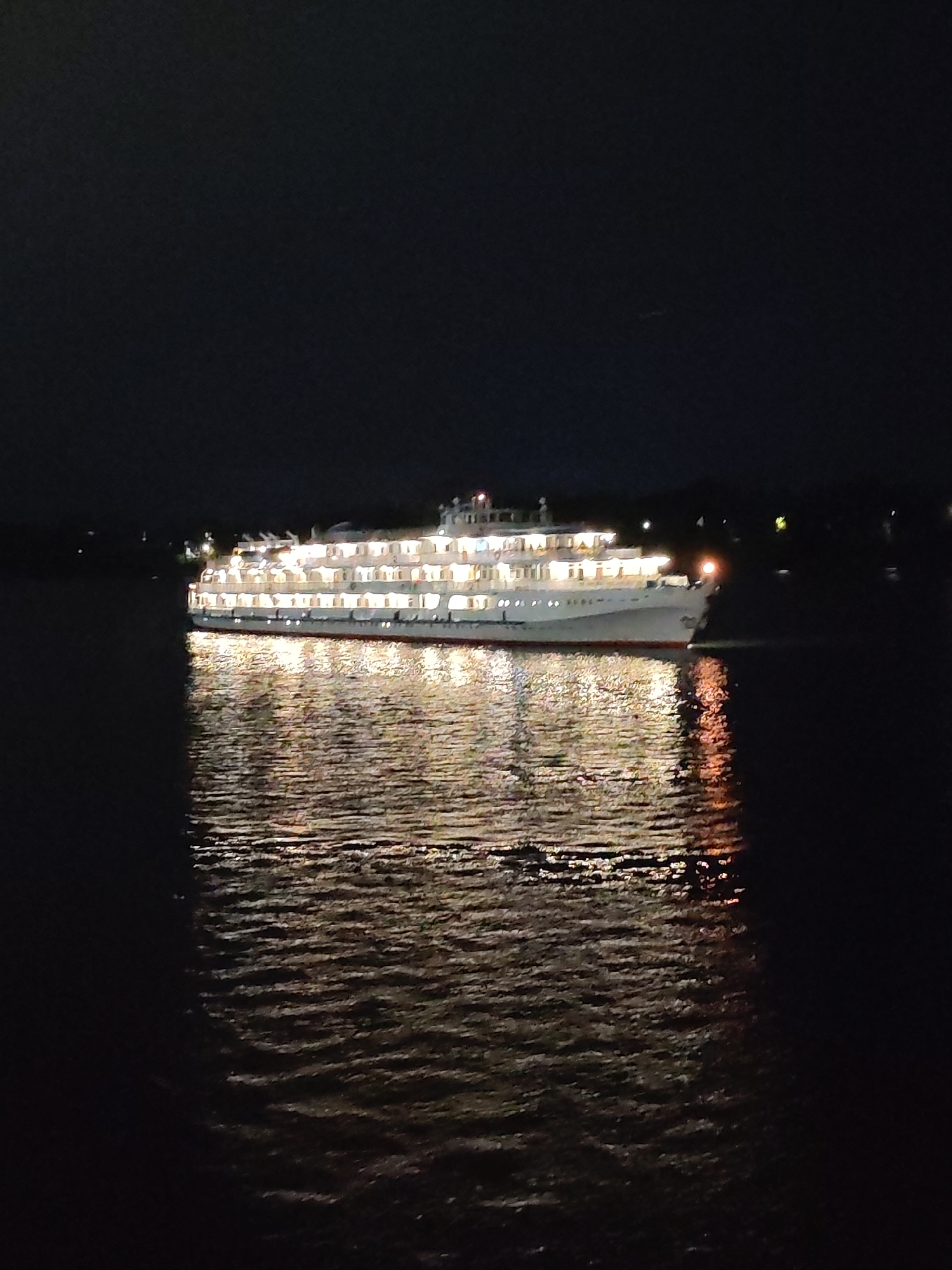 A large, multi-decked white ship is illuminated at night while floating on dark water. The ship's numerous windows glow with a warm yellow light, reflecting onto the rippling surface of the water. The night sky is dark and appears to have some distant lights visible on a dark horizon, possibly indicating land or other vessels. The water is calm with some texture from small waves.