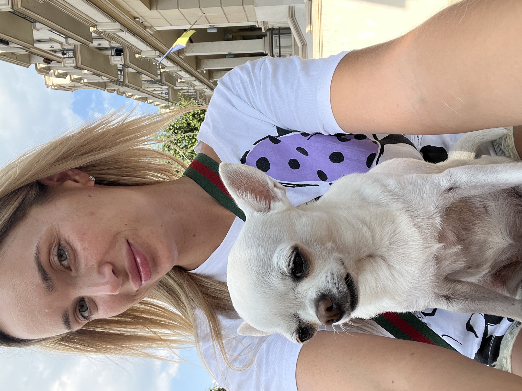 A young woman with blonde hair and green eyes is taking a selfie outdoors, holding a white chihuahua. She is wearing a white t-shirt with a purple polka dot design on it, and a strap with green and red stripes is visible across her chest. In the background, there is a building with balconies and a Ukrainian flag. The scene appears to be in a city.