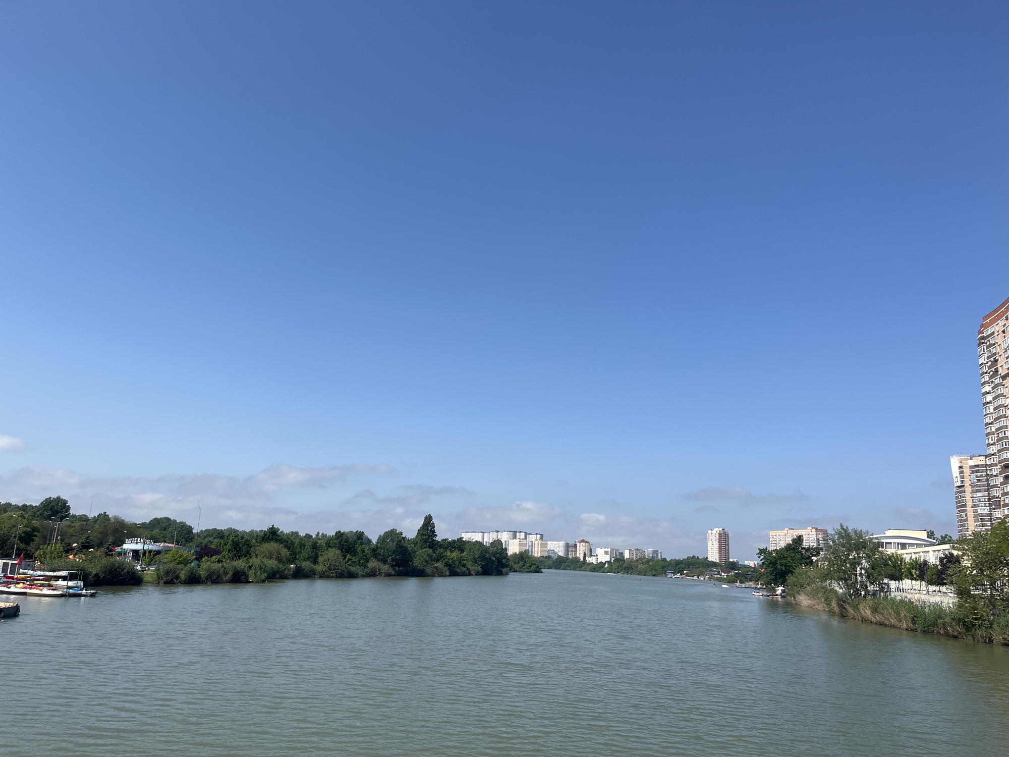 The image depicts a wide river or canal in Krasnodar, Russia, with green water under a mostly clear blue sky with a few scattered clouds. Buildings, including a tall apartment complex on the right and several lower-rise structures in the distance, line the riverbanks. Dense trees and vegetation also border the river on both sides. On the left side, there are small boats moored near a structure partially obscured by trees. The perspective is from a slightly elevated position, possibly a bridge or high bank.