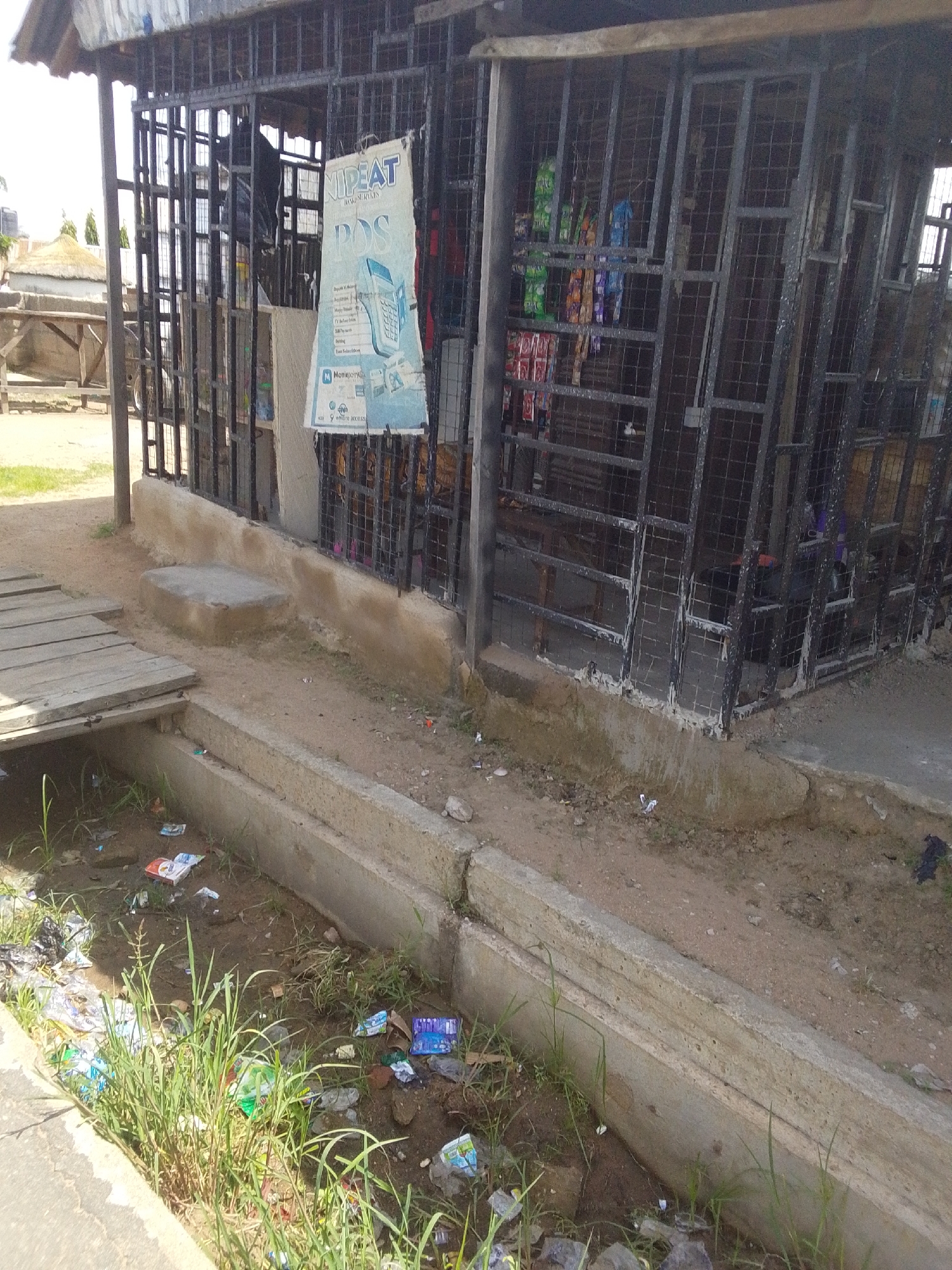 The image depicts a street scene in Jalingo, Nigeria, characterized by a small, caged shop front and a dirty drainage ditch. The shop, made of metal bars, appears to sell various goods, with some items visible through the bars, including packaged snacks and drinks. A sign advertising "NIPEAT POS" is prominently displayed on the shop's exterior. In front of the shop, a wooden walkway crosses over the drainage ditch, which is filled with stagnant water and debris. Litter, including plastic bags, wrappers, and bottles, is scattered in and around the ditch, indicating a lack of proper waste management. The ground surrounding the shop is dusty and unpaved. The overall impression is one of a typical informal market setting in a developing urban area, marked by economic activity and environmental challenges.