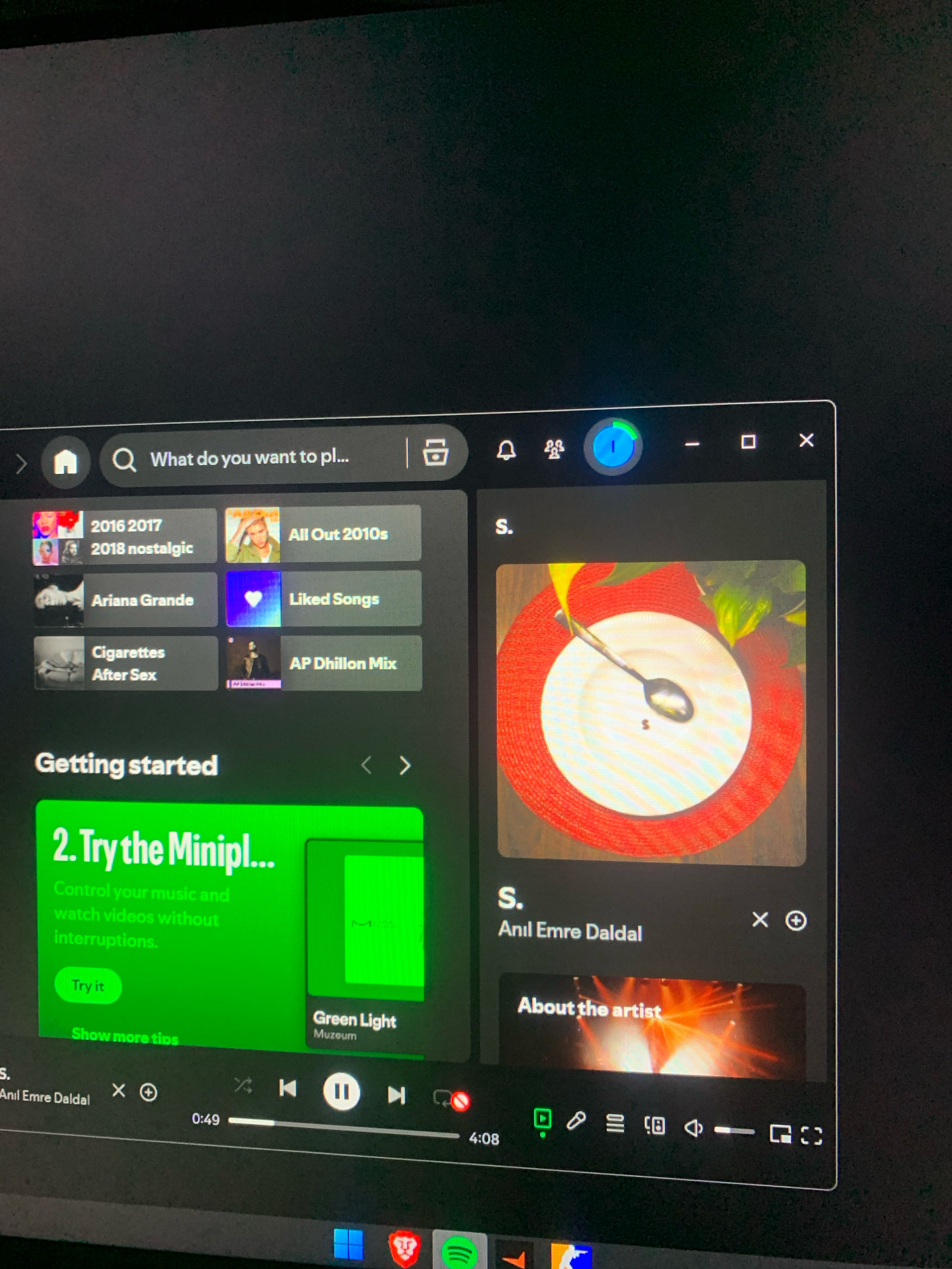 The image shows a screenshot of the Spotify desktop application. The application displays a search bar at the top, followed by a grid of album covers and playlists. Below this, there is a section titled "Getting started" with a green card featuring a "Try it" button. Further down, an artist's profile is displayed with their name and an "About the artist" section. At the bottom of the screen is the music player interface, showing playback controls, a progress bar, and the current song duration. The screenshot is taken on a computer screen, which is black and shows some reflections. There are no people, city elements, or any indicators related to Peshawar City Tehsil, Pakistan in the image.