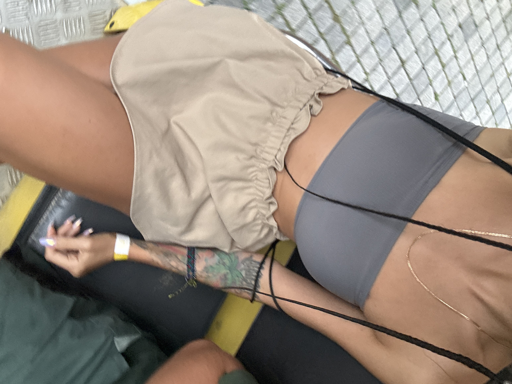 The image shows a close-up view of a person's torso and legs. The person is wearing a grey strapless top and a tan, ruffled skirt. A delicate gold chain necklace is visible around their neck. Black straps from a bag or clothing cross over the person's chest. The person has tattoos on their arm and is wearing a white and yellow wristband. Their fingernails are painted with a holographic design. The background is blurred but appears to be an outdoor setting with patterned paving stones and a dark surface, possibly seating. The location is described as Tây Hoa Lư Ward, Vietnam.