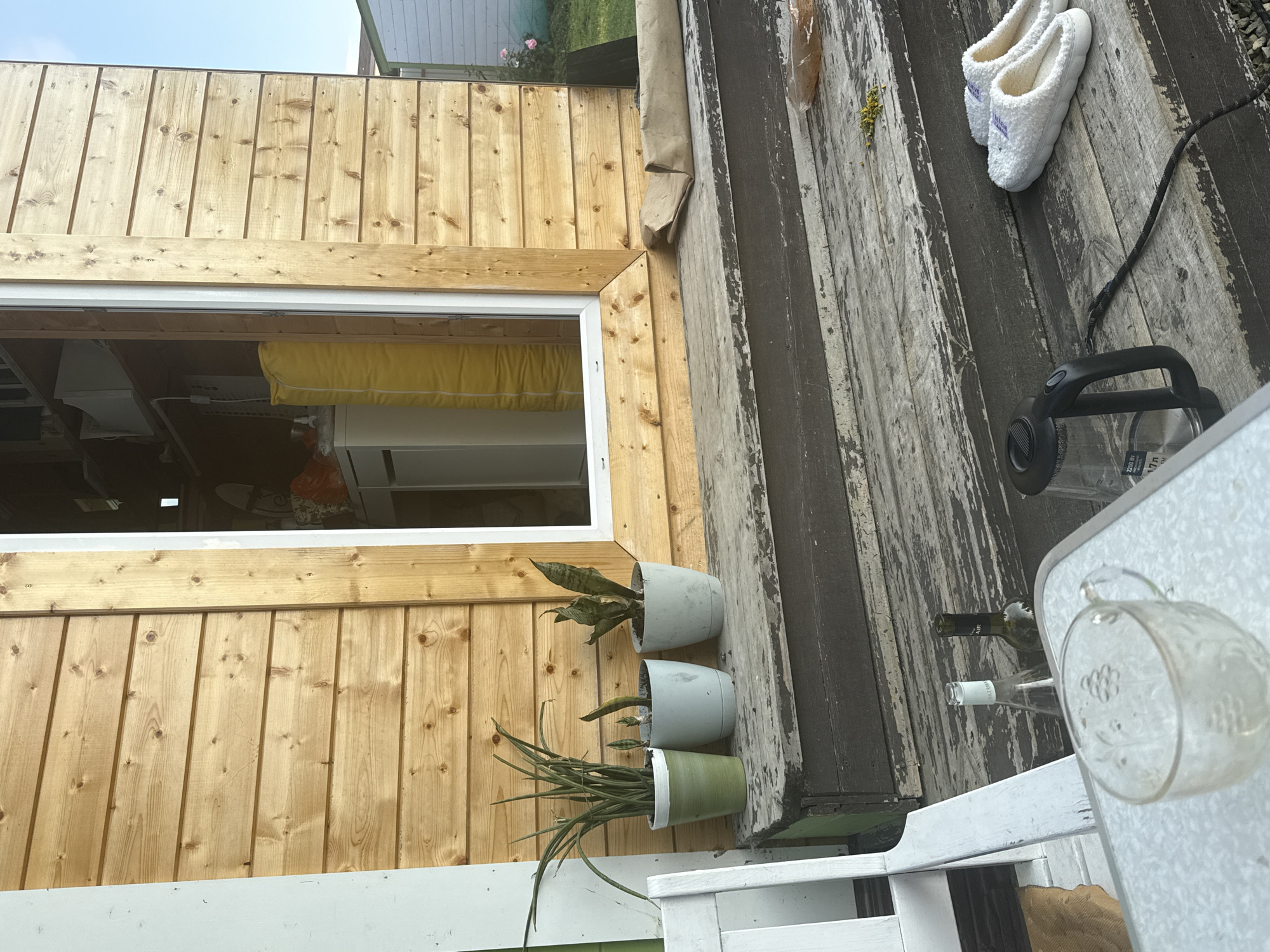 A close-up shot captures a wooden exterior of a structure with horizontal planks, featuring a doorway with a white frame. Three potted plants, identifiable as snake plants (Sansevieria trifasciata), are arranged on a weathered wooden surface in front of the structure. The plants are in light-colored pots, with one pot being green.
In the foreground, wooden steps lead up to the structure. The steps are dark and show signs of wear and peeling paint. On the steps, a black electric kettle with a cord, a pair of white fluffy slippers, and scattered yellow flower petals are visible. Further to the left, on a white table, a clear glass cup with an embossed floral pattern is present, along with two glass bottles - one appears to be a wine bottle, and the other is a clear glass bottle with a white label.
The scene suggests an outdoor setting, possibly a porch or backyard area of a small house or cabin. The context of Lyady, Russia, implies this is likely a domestic or rural location. There are no people visible in the image. The lighting indicates it is daytime, with sunlight casting shadows and highlighting the textures of the wood and objects. The overall impression is one of a lived-in, perhaps slightly rustic, outdoor space.