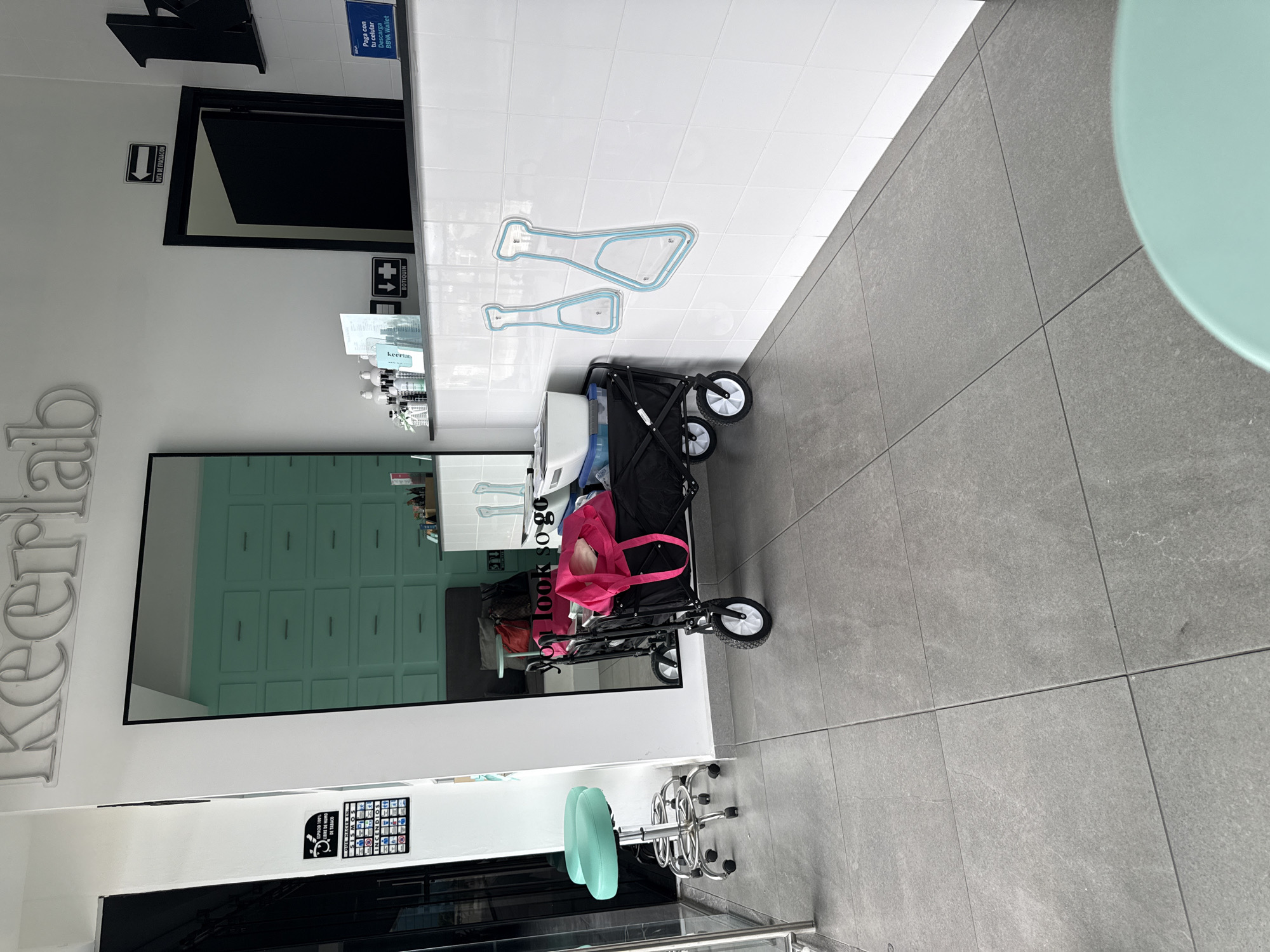 This is an interior shot of a business, likely a spa or salon called "Keerlab," based on the signage visible. The image is rotated, showing the floor on the right and the ceiling on the left. The room has a minimalist aesthetic with white walls and grey tiled floors. On the left is a mirrored wall reflecting a storage cabinet with light green doors. In front of the mirror is a black cart with white trim, carrying various items including a white container, a blue item, and a pink bag. Behind the cart is an adjustable stool with a green seat and chrome base. The wall on the opposite side is white with two embedded clear, outlined shapes on it. Signs are visible on the wall, including safety and informational signage. The room is brightly lit. The location is indicated by the OCR to be in San Bernardino Tlaxcalancingo, Mexico.