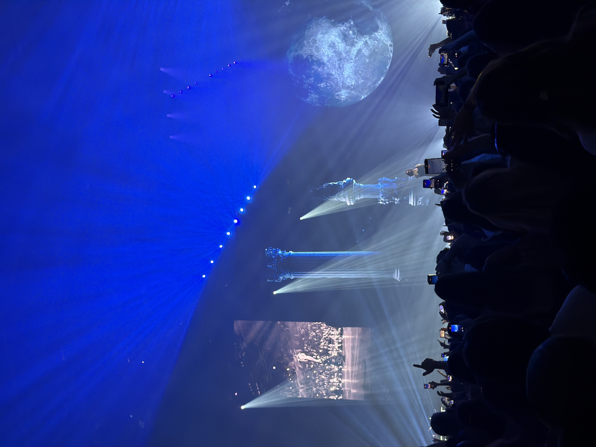 The image depicts a concert or live performance, possibly in Sofia, Bulgaria, although no specific landmarks confirming the location are evident. The audience is visible on the right side of the frame, holding up their phones to record the event. The stage setup includes a large, spherical screen displaying an image of the moon. Stage lighting is prominent, featuring blue lights and spotlights directed towards the performers or stage elements. There are also screens displaying images. The overall ambiance suggests a high-tech, visually immersive concert experience.