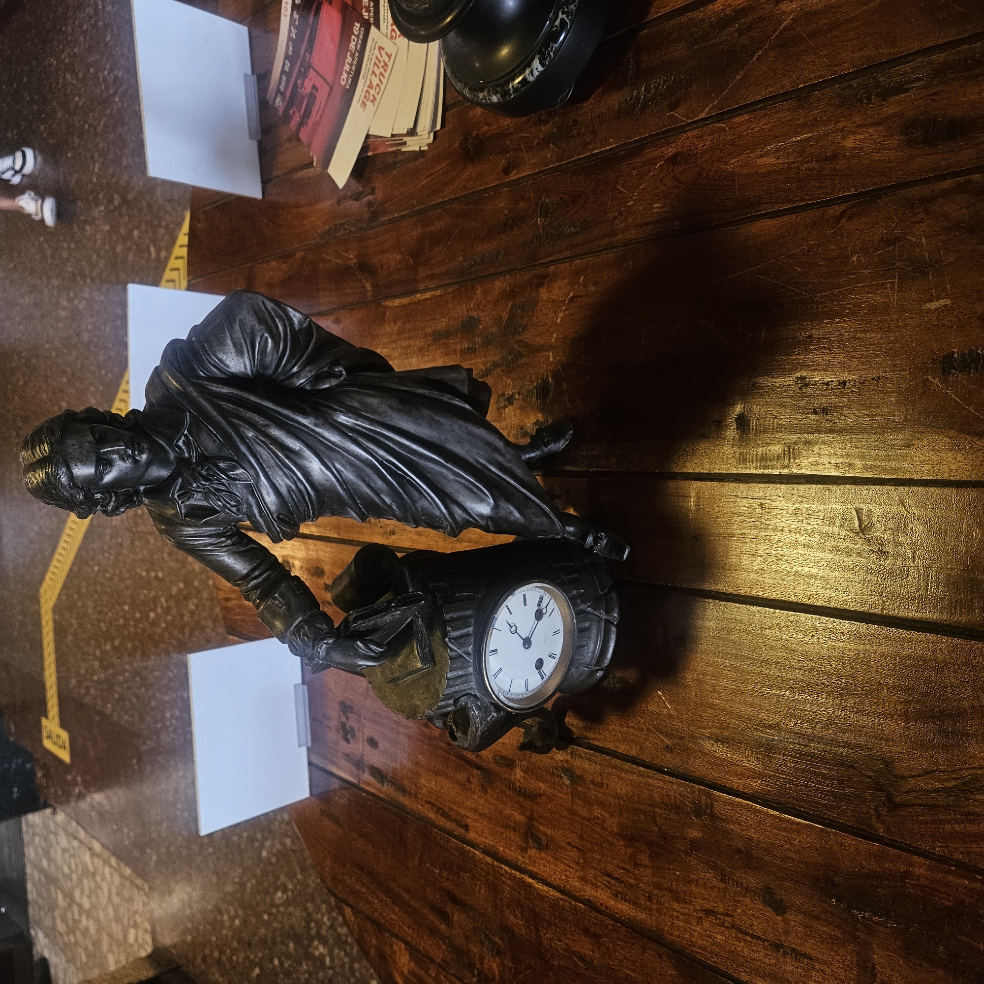An outdoor, eye-level, medium shot depicts a bronze statue of a man holding a book and scroll, standing next to an antique clock on a wooden table. The statue's dark bronze finish contrasts with the table's rich, polished wood grain. The clock, also appearing to be made of bronze, features a white face with Roman numerals and black hands, indicating a time of approximately 4:10. In the background, white signs are visible, and a yellow arrow painted on the floor points to the right. Scattered on the table are papers with red and white lettering, partially obscured by the statue and clock. The overall impression is one of a curated display, possibly in an open-air market or exhibition space. The specific location in Almadrones, Spain, is not discernible from the visual information provided in the image.