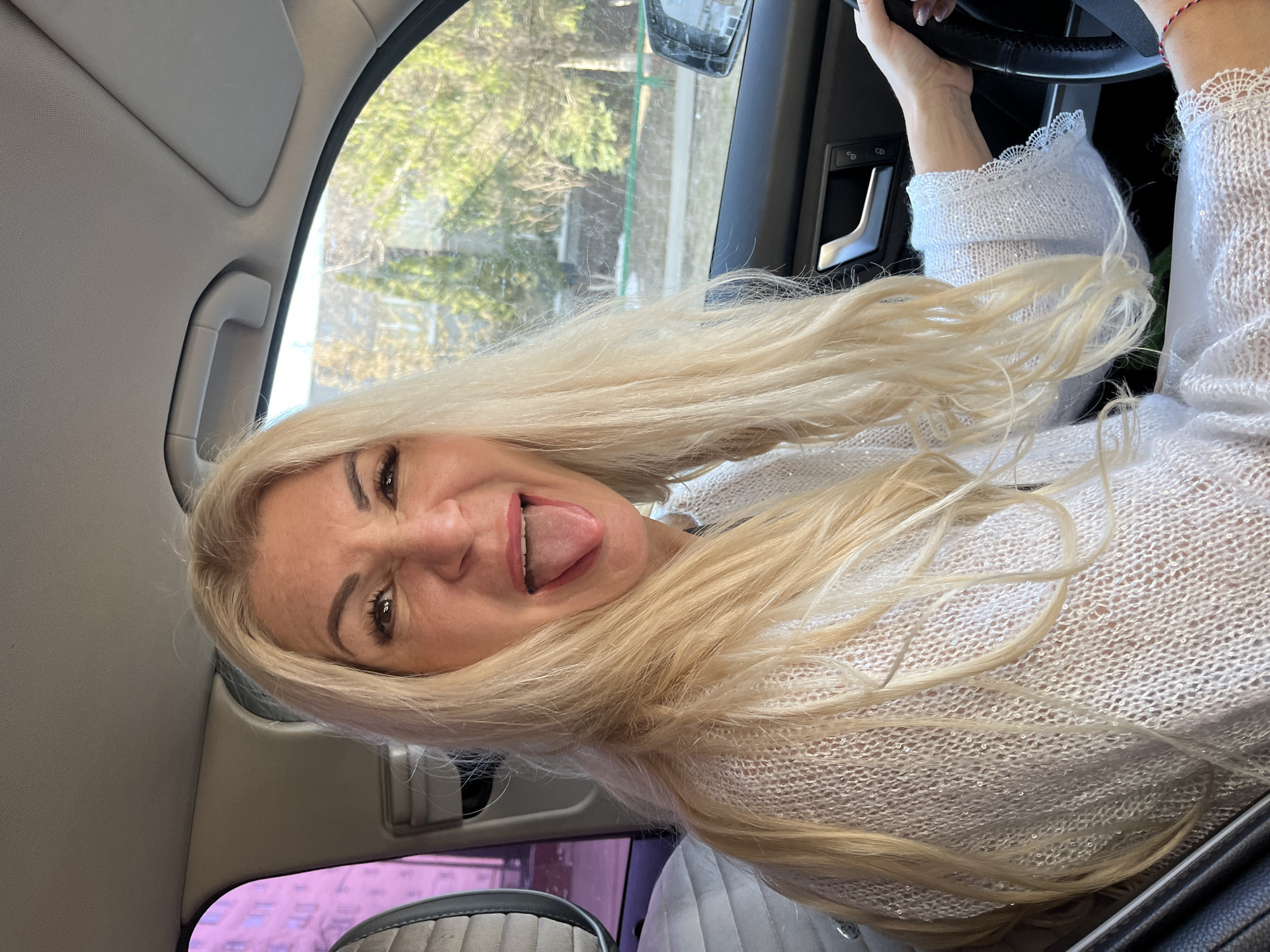 A woman with long blonde hair is inside a car, possibly a taxi, sticking out her tongue. She is wearing a white knitted sweater. Her hands are visible on the steering wheel. The car is moving. Through the window, there is a blurred view of an urban environment, possibly Nizhny Novgorod, Russia. The time of day appears to be daytime.