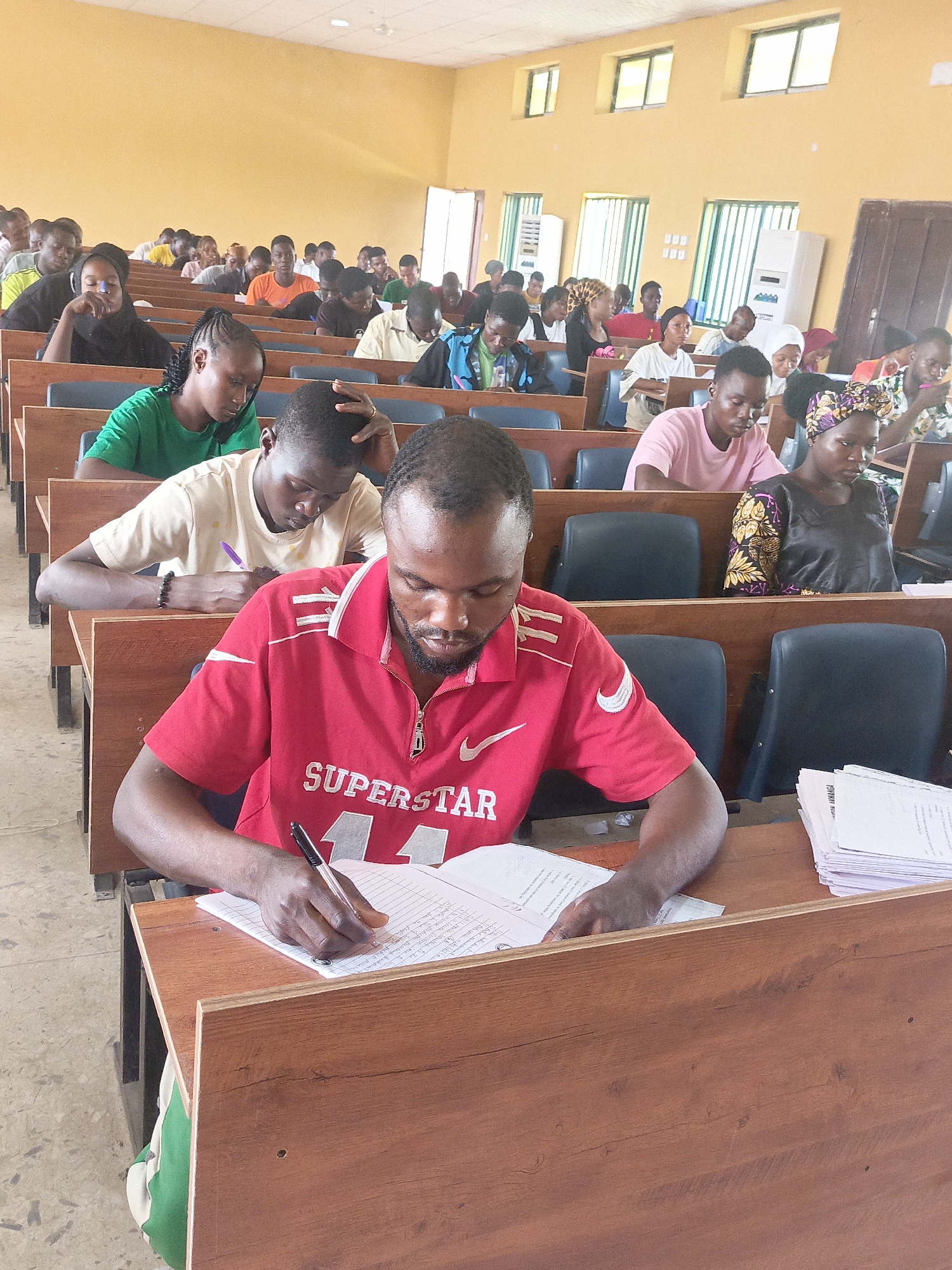This image depicts a bustling classroom or lecture hall in Akwanga, Nigeria, filled with numerous young adult students. The setting features rows of wooden desks with attached black chairs, arranged in an academic environment. The walls are a light yellow or ochre color, and several windows with vertical security grilles are visible, allowing natural light to enter. Overhead fluorescent lights supplement the illumination.
The primary activity taking place is academic work, with most students focused on writing or studying. In the immediate foreground, a man wearing a red polo shirt with "SUPERSTAR" and a Nike swoosh logo is seen diligently writing on a notebook with a black pen. Handwritten text is visible on his paper. To his right, a stack of white papers rests on the desk, one sheet partially revealing the text "FOWMANGA" and "QUESTION."
Behind him, other students are equally engaged. A woman in a green t-shirt is writing, while next to her, a man in a light-colored t-shirt is also writing, with one hand resting on his head in a gesture of concentration. Further back, numerous other students, both male and female, are seated, absorbed in their tasks. Some wear headwraps, while others are in casual attire like t-shirts. The expressions on their faces generally convey focus and seriousness, indicative of an exam or intensive study session. The scene appears to take place during daylight hours.