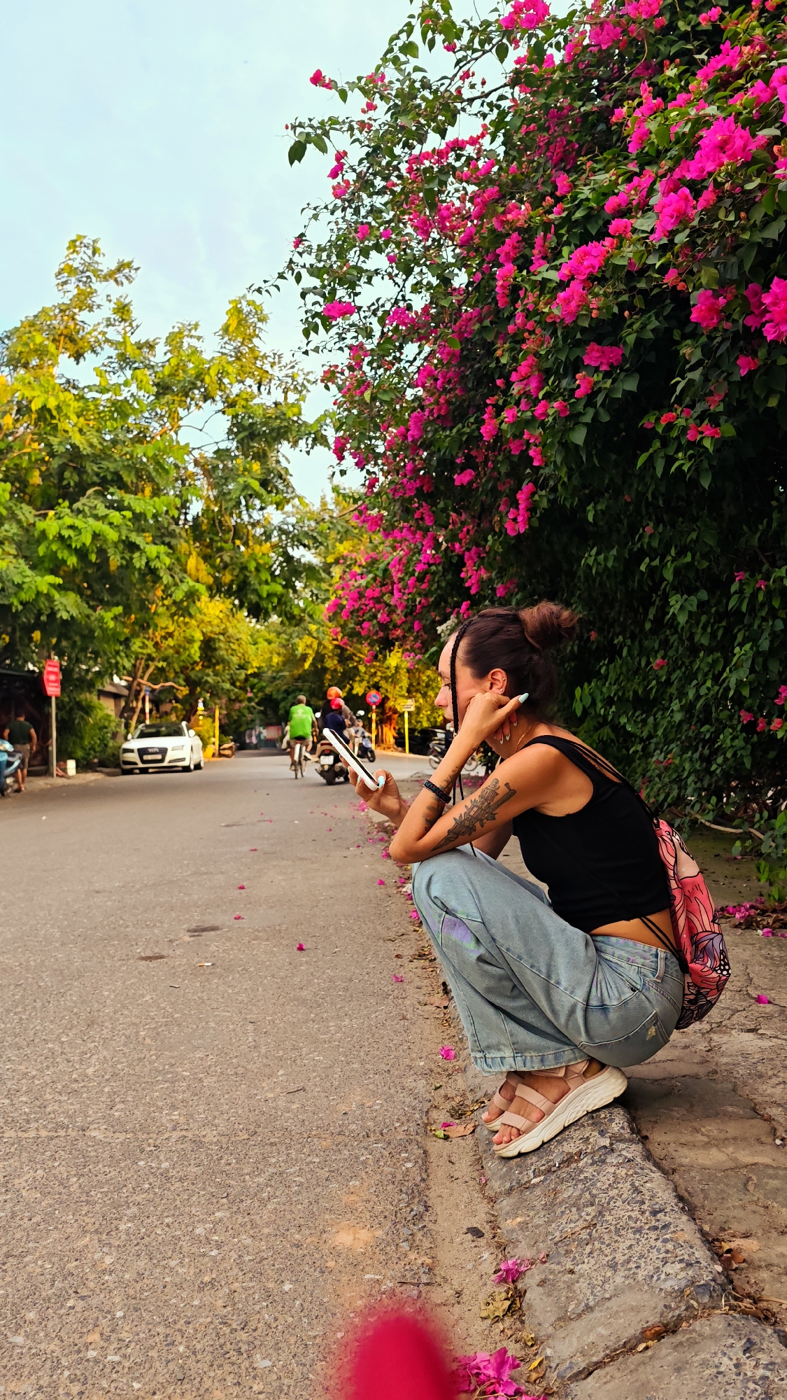 An outdoor, eye-level shot shows a young woman crouched on a sidewalk in Hanoi, Vietnam, looking at her phone. She is wearing a black tank top, light blue jeans, and light pink sandals. A pink backpack with a butterfly pattern is visible on her back. Her right arm is bent, supporting her head as she looks at the phone. Tattoos are visible on her left arm. She has dark hair pulled up in a bun with a braid.
The sidewalk is a concrete curb along an asphalt road. Pink flower petals are scattered on the sidewalk and road. Behind the woman, a vibrant bougainvillea bush with bright pink flowers forms a dense hedge. To the left of the road, trees with green foliage line the street. Several vehicles are on the road, including a white car in the middle ground, and a person on a bicycle wearing a green shirt. The sky is a light blue with some clouds. The overall lighting suggests late afternoon or early evening.
