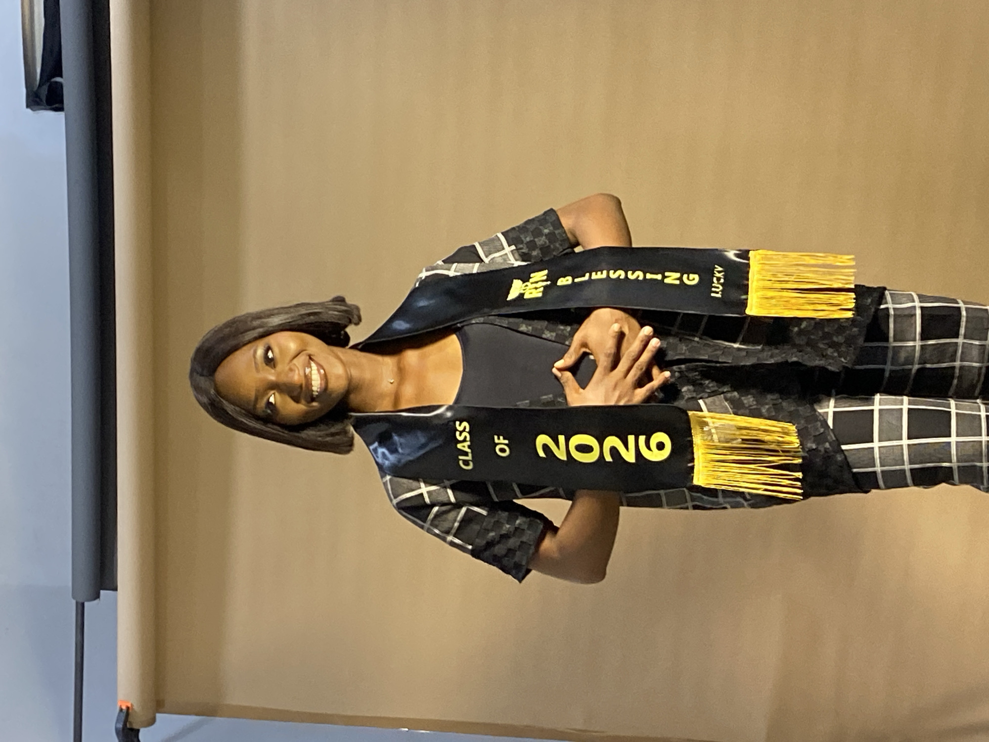 A dark-skinned woman is centered in the image, smiling and wearing a black sash with "CLASS OF 2026" printed on one side and "BLESSING LUCKY" on the other, both in yellow lettering. The sash has yellow fringe. She is wearing a black and white checkered patterned top over a black top. The background is a plain tan or light brown color. A section of a black frame is visible on the left side of the image. No city or country is visible, so no statement can be made of Nigeria.