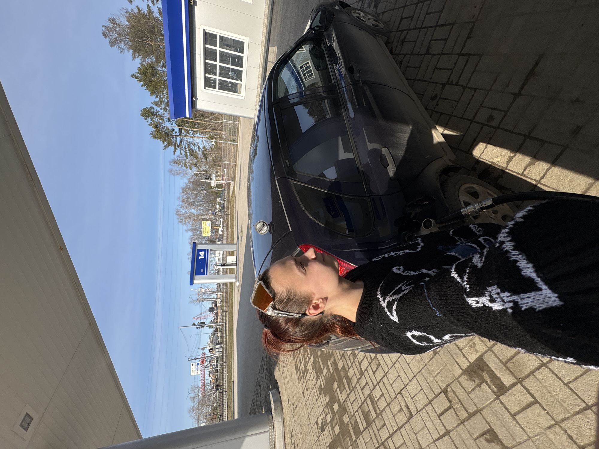 A young woman is filling a dark-colored car with gasoline at a fuel station in Chaikovsky, Russia. The woman has reddish-brown hair pulled back in a low ponytail and is wearing yellow-tinted sunglasses and a black and white patterned sweater. The car is parked on a brick-paved surface near a fuel pump. In the background, there's a building with a window, trees, and a sign for fuel pump number 14. The sky is clear and blue.