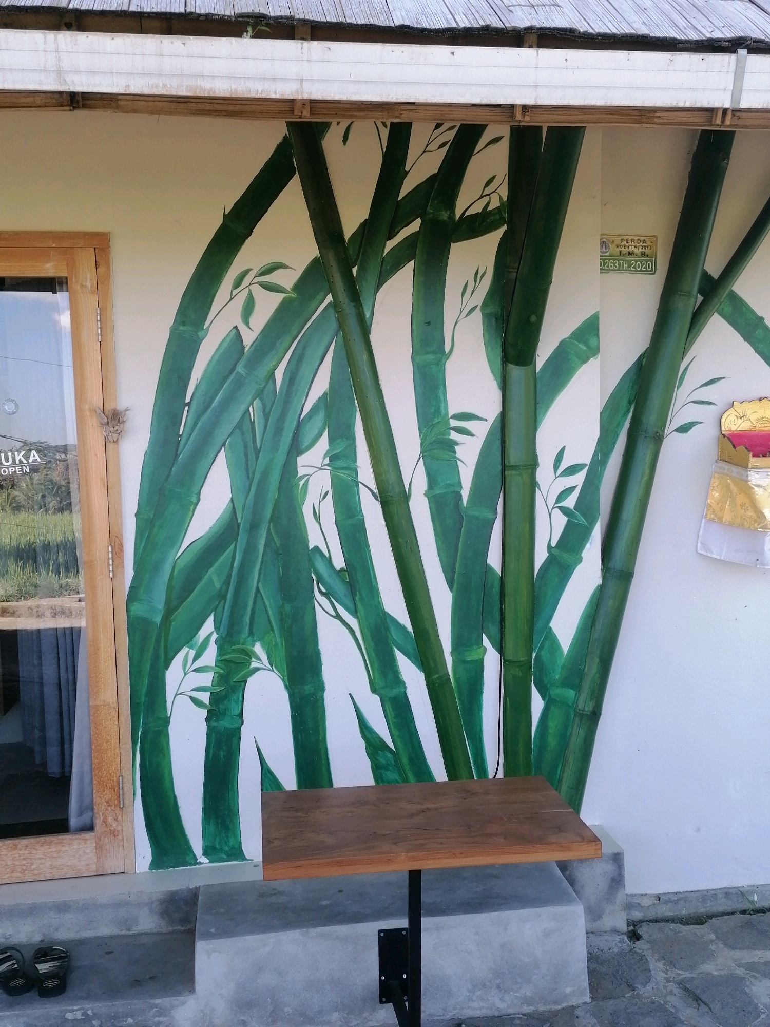 The image depicts a section of a wall adorned with a vibrant mural of green bamboo stalks, painted in a realistic style with subtle shading. The bamboo extends from the lower left to the upper right of the frame, with a few delicate green leaves branching off. To the left, a wooden framed glass door is visible, partially open, revealing a glimpse of what appears to be an indoor space with curtains. A small, light brown butterfly is perched on the wooden door frame.
In front of the mural, a small, dark brown wooden table with a black metal stand is positioned on a gray concrete platform, which is elevated from the ground. Below the platform, on the gray concrete steps leading down, a pair of black flip-flops are placed.
On the right side of the mural, a small, decorative religious offering, likely Balinese in style, is attached to the wall. It features gold and white fabric with a hint of red and pink, topped with a golden element. A small rectangular sign with text is also affixed to the wall near the top right of the bamboo mural.
The background of the scene suggests an outdoor setting, possibly a cafe or shop entrance, with hints of greenery and outdoor flooring visible. The overall impression is one of a serene and naturalistic aesthetic integrated into a commercial or hospitality space. The location is identified as the city of Ubud District, Indonesia.