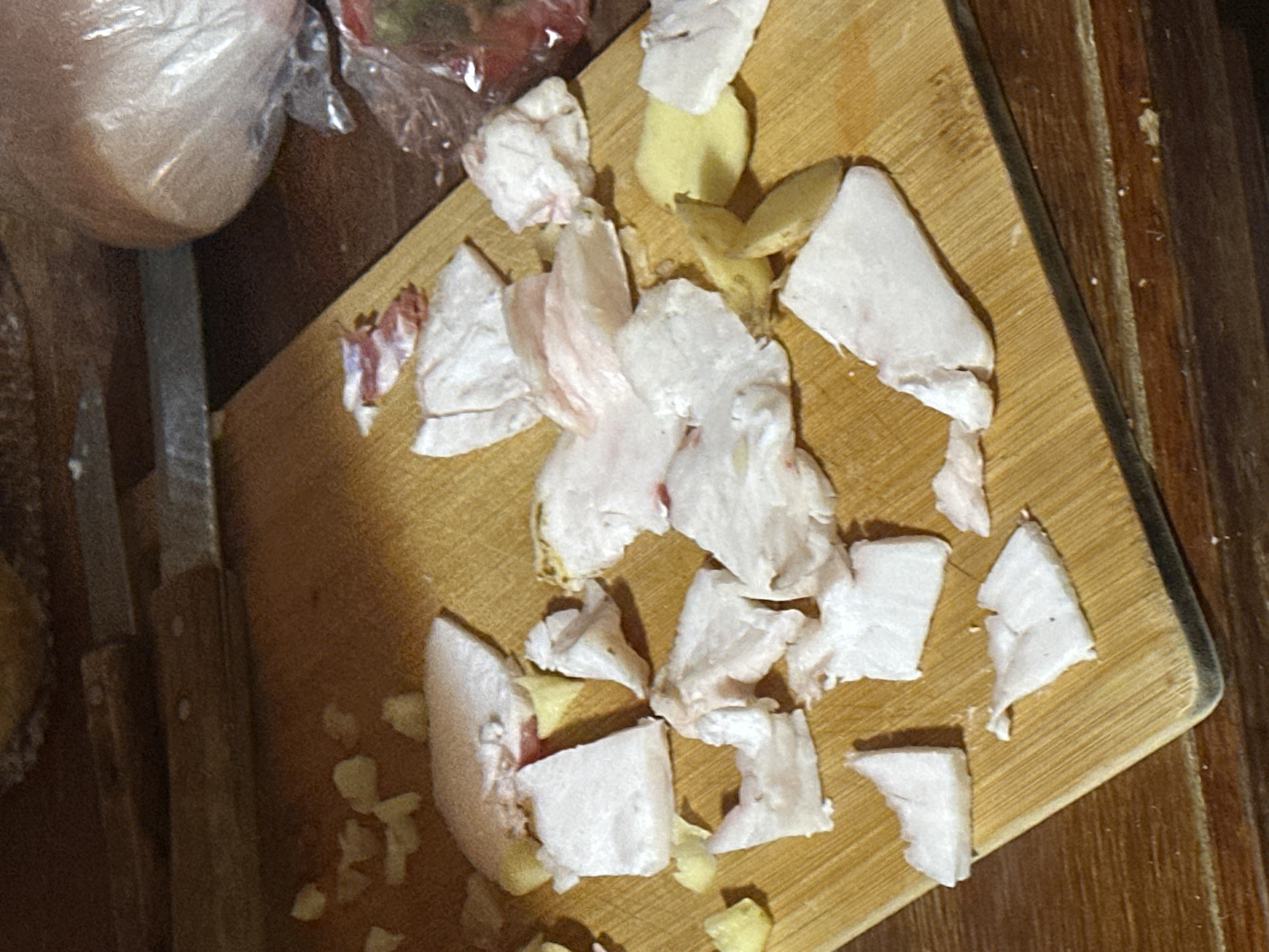 The image depicts a cutting board on a wooden table, strewn with various pieces of white fat, likely pork fat, some with pinkish hues. There are also a few slices of yellow potato and some small, irregular white fragments scattered across the board. In the background, out of focus, is a knife and a plastic-wrapped object, possibly food or produce. The lighting is dim, casting shadows and highlighting the texture of the fat. The scene appears to be a domestic kitchen setting. The image is a close-up shot of the cutting board and its contents, with no visible indication of its location in Arkhyz, Russia.