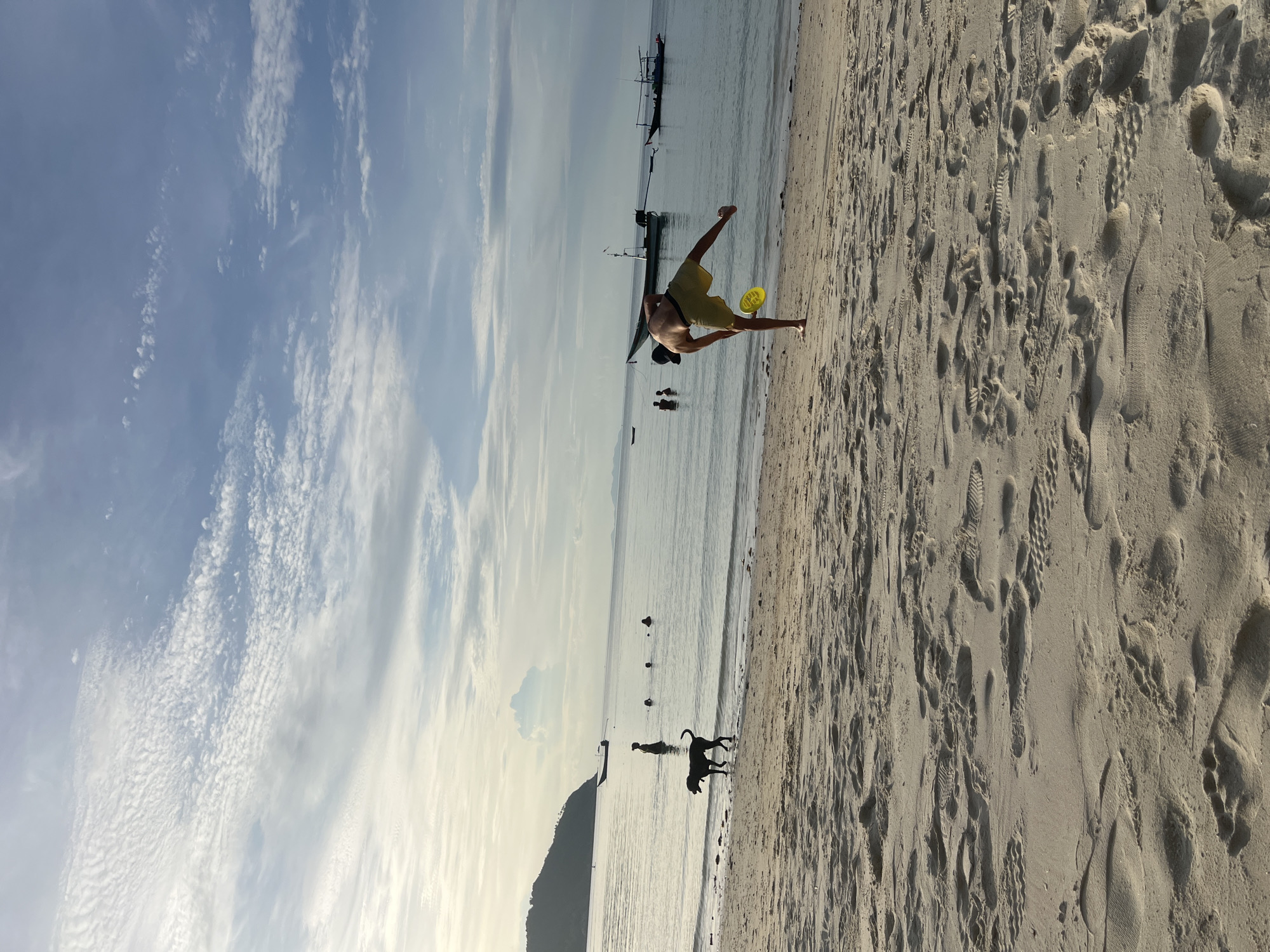 An outdoor, eye-level, full shot captures a beach scene on a cloudy day. In the foreground, the sand is covered with footprints. A person wearing yellow shorts and a black hat is in the middle of the frame, mid-action, throwing a yellow frisbee. The person is facing away from the camera and is balanced on one leg with the other leg extended. In the background, the calm ocean water meets the sandy shore. Several people are in the water, and some are further out. A black dog stands on the beach near the water's edge, facing left. Boats are visible on the water, and a hill or island can be seen in the distance. The sky is filled with scattered white clouds.