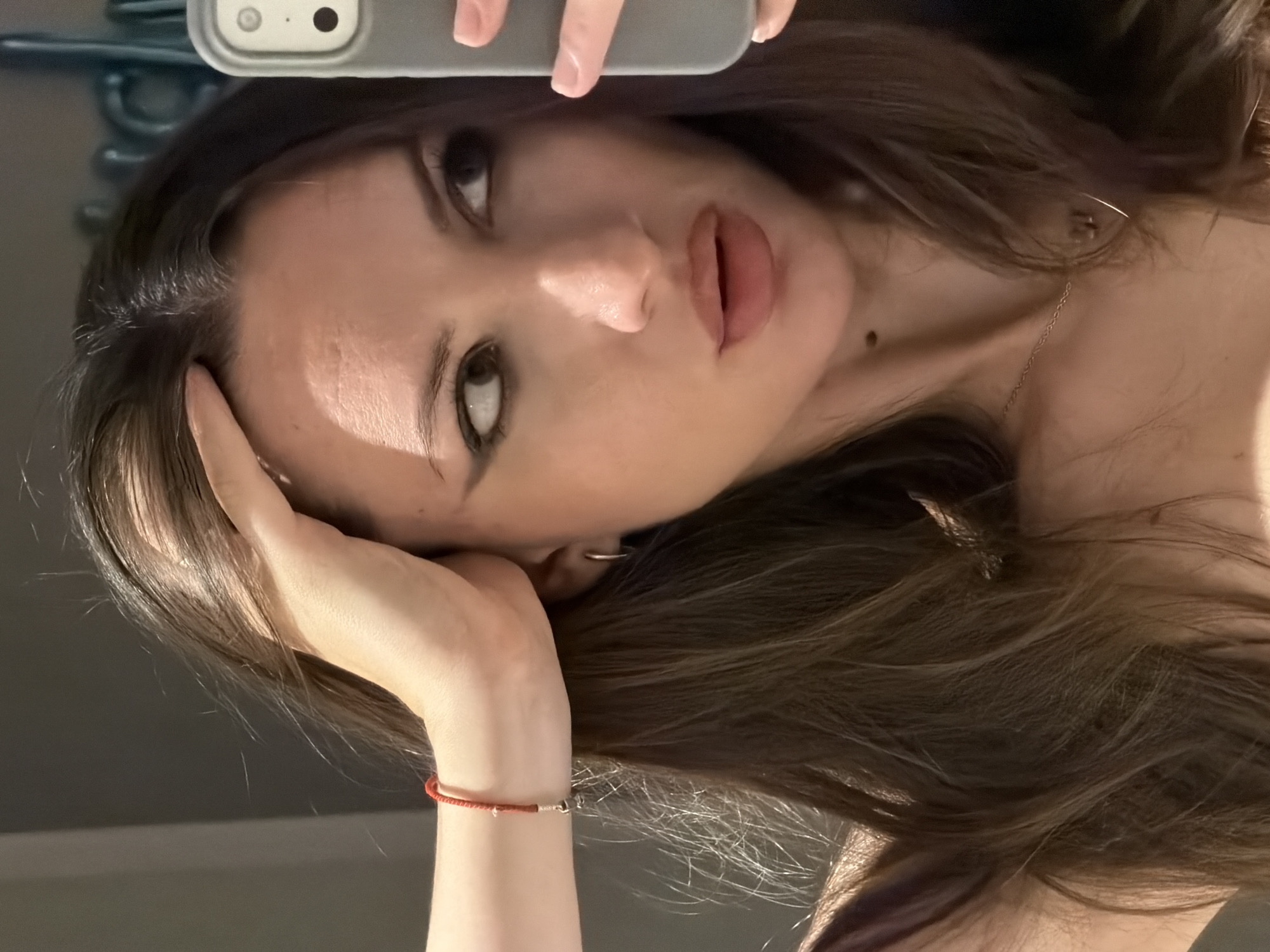A woman with long, dark brown hair is taking a selfie in a mirror. She is wearing a dark-colored top and a thin silver necklace. A red string bracelet with a small silver charm is visible on her left wrist. Her makeup includes dark eyeliner and lip gloss. The background is a blurred interior, possibly a bathroom, with a grey wall and a hint of what appears to be a blue-tinted mirror or decorative element. The scene is not identifiable as being in Sharm El Sheikh, Egypt.