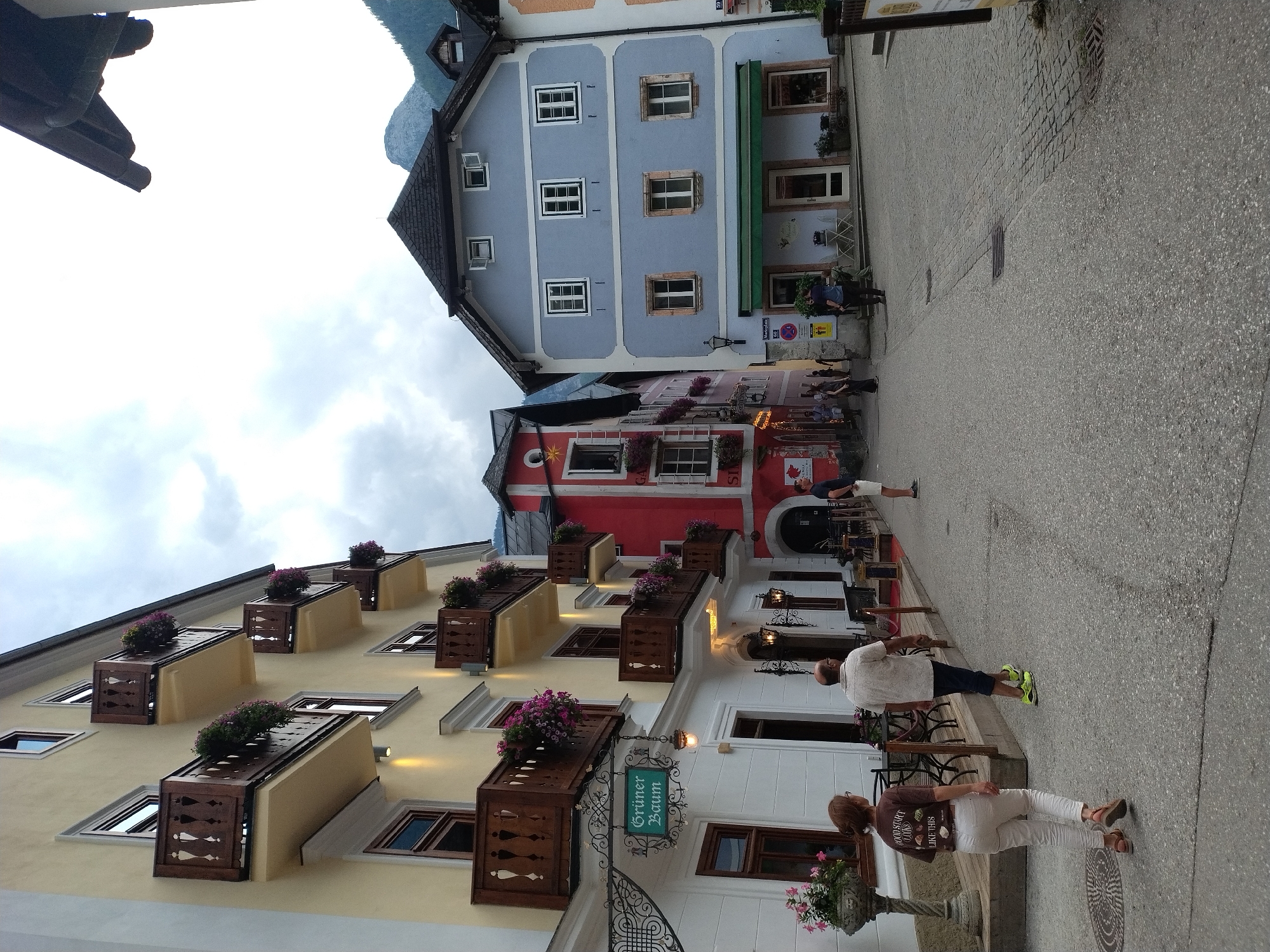 The image shows a street scene in Hallstatt, Austria, with several buildings lining a cobblestone street. On the left, a yellow building with multiple balconies decorated with wooden railings and potted flowers extends upwards. A sign reading "Grüner Baum" hangs from the building. Next to it is a red building, also with decorative balconies and flower boxes. On the right side of the street, a light blue building features a dark roof and several windows. Green awnings are visible above some of the ground-floor shops. Several people are walking along the street. A woman in white pants and a brown shirt is walking away from the camera on the left. A man in a light-colored shirt and dark shorts is walking ahead of her. Further down the street, other individuals are seen walking or standing near the buildings. The sky is overcast. The overall scene depicts a charming European town with traditional architecture and a pedestrian-friendly atmosphere.