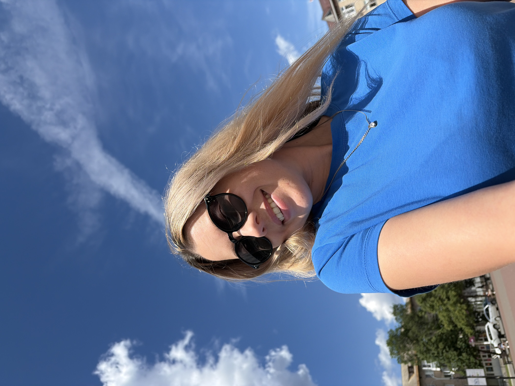 A young woman with blonde hair and sunglasses smiles at the camera while wearing a blue t-shirt and a necklace. The background shows a bright blue sky with white clouds and a cityscape with buildings, trees, and cars. The woman is positioned in the foreground, with her head tilted slightly to the left. The image appears to be a selfie taken outdoors on a sunny day in the city of Chaikovsky, Russia.