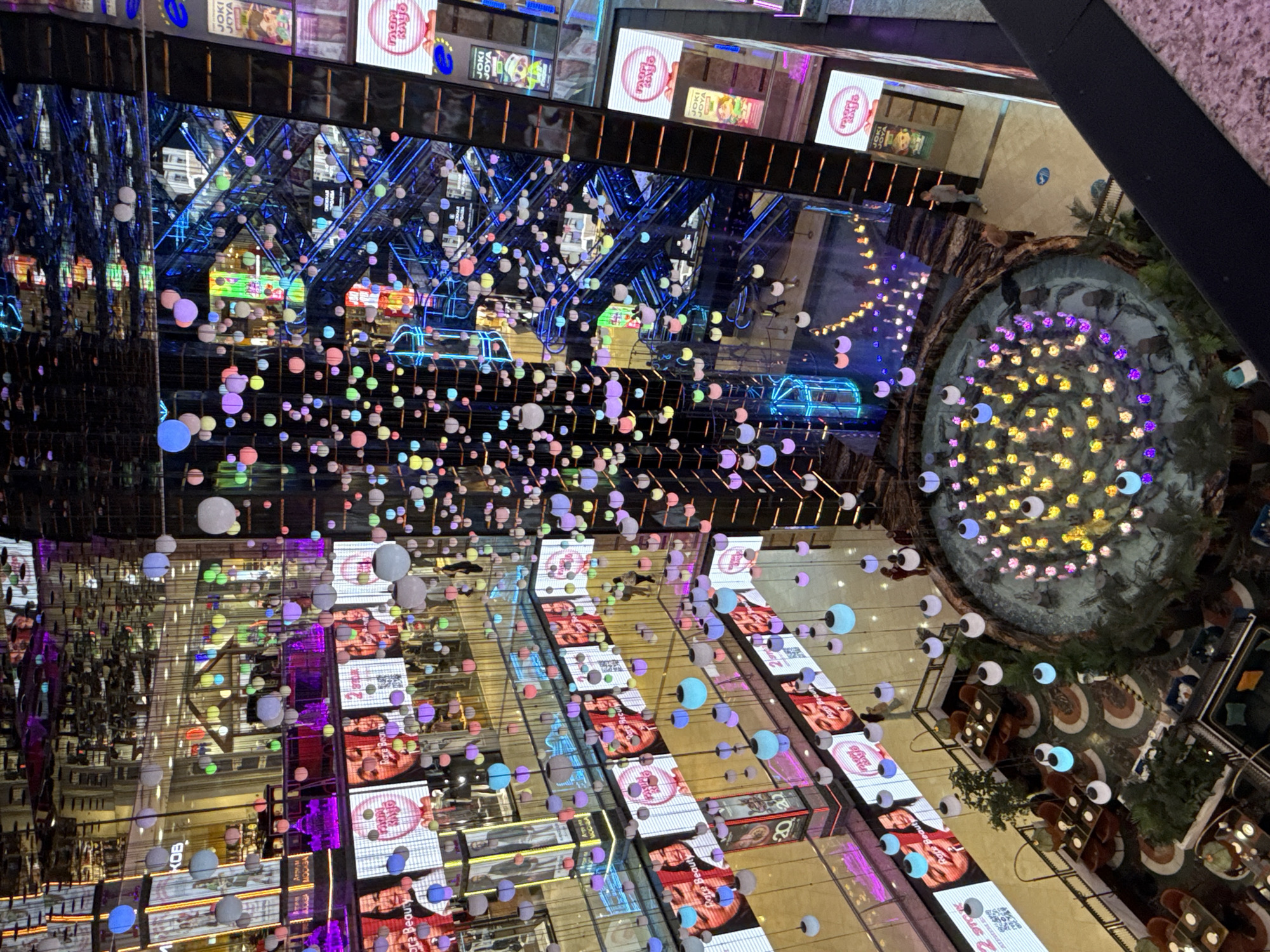 The image captures a multi-level interior view of a shopping mall, likely in Moscow, Russia, characterized by its vibrant and modern aesthetic. Suspended from the ceiling are numerous colorful spherical lights in various shades of blue, pink, yellow, and white, creating a festive and dynamic atmosphere. These lights are strung at different heights, adding a sense of depth and visual interest to the scene.
The mall's architecture features several levels, connected by escalators and walkways. The escalators, illuminated with bright blue neon lights, are a prominent feature, drawing the eye upwards and suggesting a busy environment. Large digital screens are mounted on the walls of different levels, displaying advertisements and images. Some screens feature portraits of people, while others showcase product promotions.
In the foreground, a circular fountain with colorful underwater lights is visible, adding a serene element to the bustling mall. The fountain area is surrounded by decorative stones and lush green plants, enhancing the natural ambiance. Nearby, several seating areas with tables and chairs are arranged, indicating dining or relaxation spaces within the mall.
Several individuals are present in the scene, though their interactions are not clearly discernible. They are seen walking on the walkways and escalators, suggesting normal mall activity. The overall environment is one of commercial activity, entertainment, and modern design, with a focus on visual stimulation through lighting and digital displays. The mirrored ceilings and reflective surfaces further amplify the sense of space and light, creating a visually rich and complex environment.