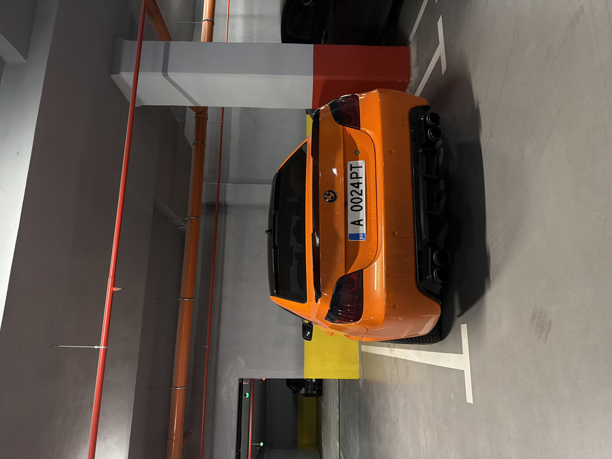 The image displays the rear of a vibrant, metallic orange car, likely a BMW X6 M, parked in an indoor concrete parking garage. The car is positioned with its rear facing the viewer, slightly angled, showcasing its quad black exhaust pipes and dark taillights. The visible license plate reads "A 0024 PT," featuring the EU flag and "RO" for Romania, indicating the car's registration.
The setting is a typical multi-story or underground parking facility. Grey concrete walls and pillars dominate the background, accented by a horizontal beam painted in sections of white and red. Several red and orange pipes, secured with metal clamps, run vertically and horizontally, suggesting utility or fire suppression systems. White painted lines on the grey concrete floor delineate the parking space. A yellow block or painted area is visible on the ground behind the car. Another dark-colored vehicle is partially visible further back in the garage. The lighting is artificial, characteristic of an indoor environment. No individuals are present in the scene. The image itself is rotated 90 degrees clockwise from its natural orientation.