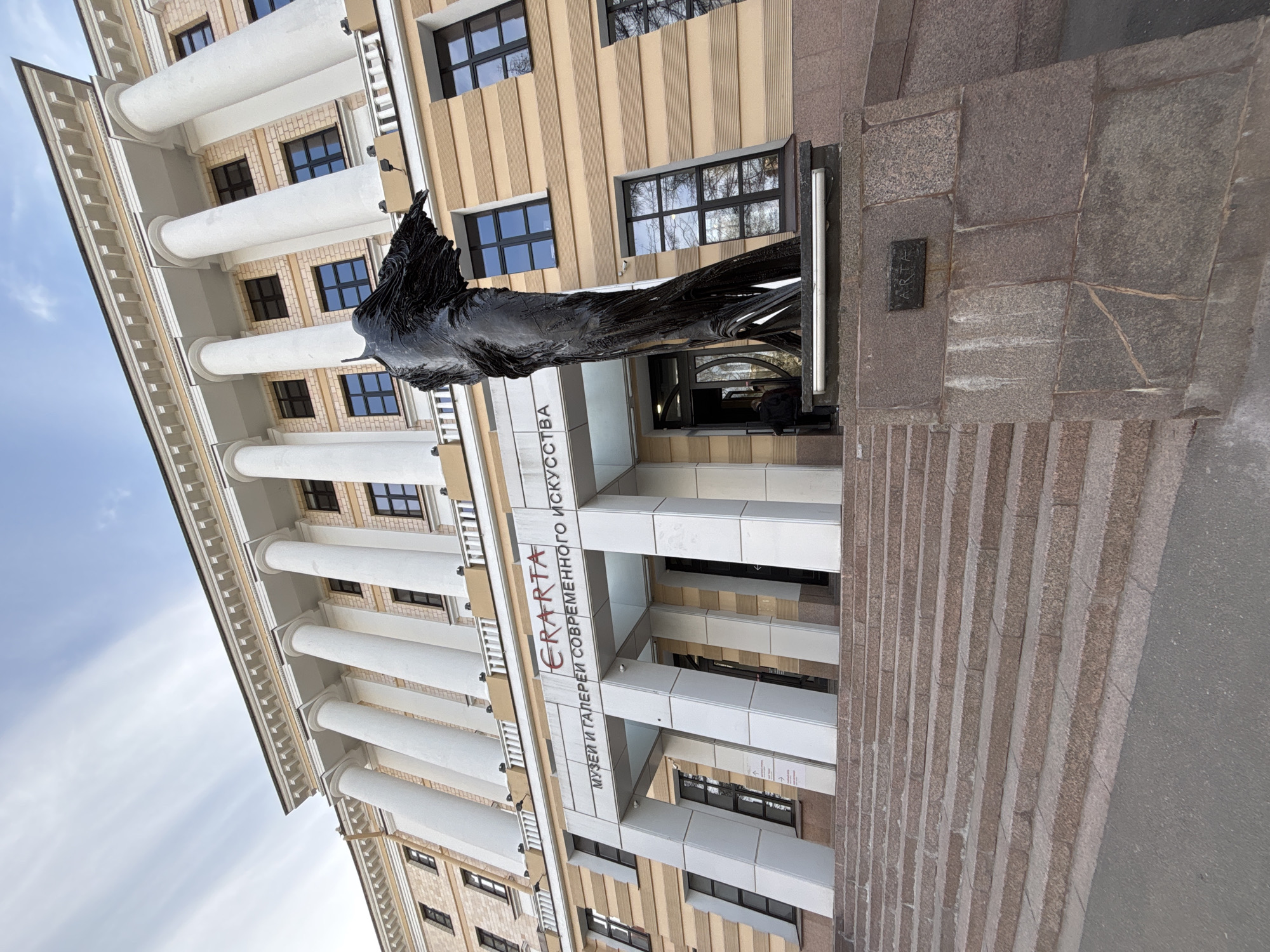 The image depicts the ERARTA Museum of Contemporary Art in Saint Petersburg, Russia. The building has a classical architectural style, with a façade featuring prominent white columns and a beige and brown striped exterior. A dark, abstract sculpture of a winged figure is prominently displayed at the entrance. The museum's name, "ERARTA," is inscribed on the building in red Cyrillic letters. The entrance area is lined with granite steps and features a modern design. The sky above is partially visible, exhibiting a light blue hue with wisps of clouds.
