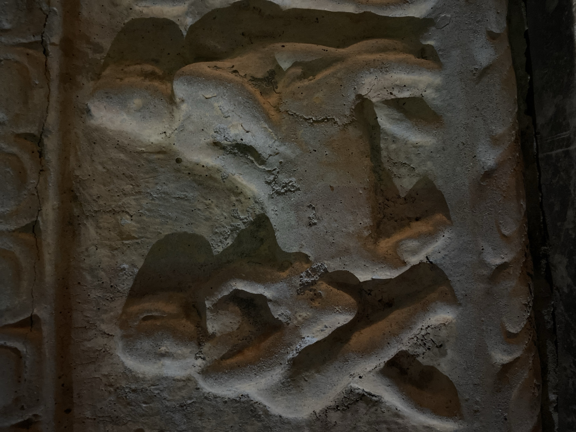 The image depicts a weathered, possibly ancient stone carving or relief. The carving features human-like figures in what appears to be a wrestling or fighting pose. The details of the figures are worn, making it difficult to discern specific features or clothing. The stone itself is textured and shows signs of age and wear, possibly indicating an outdoor location exposed to the elements. The photograph's dark and uneven lighting accentuates the carving's contours and adds to the image's antiquity. The location is Budva, Montenegro.