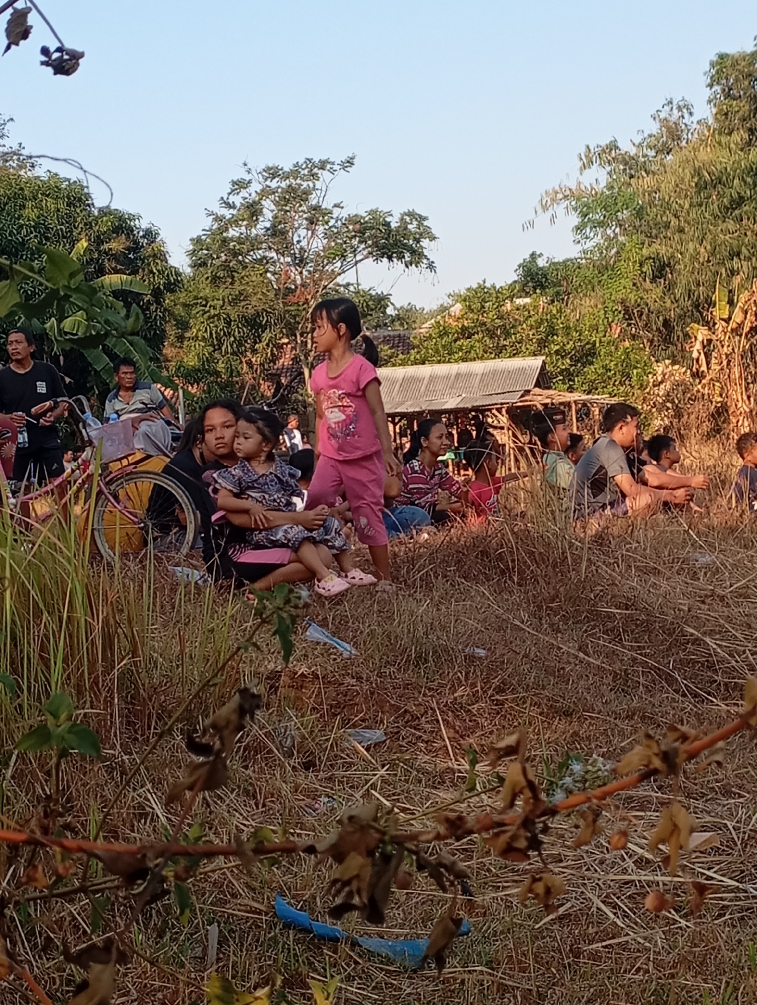 A group of people, predominantly children, are gathered outdoors in what appears to be a rural area. The scene is characterized by dry, tall grass and a clear sky. In the foreground, a woman is holding a young child. To the left, a bicycle with a yellow frame and pink wheel rims is propped up. Further back, other individuals, including adults and children, are seated or standing, observing something out of frame. Some are gathered near a simple, open-sided structure with a corrugated metal roof. The environment suggests a warm, possibly arid climate. The location is identified as Kecamatan Haurgeulis, Indonesia.