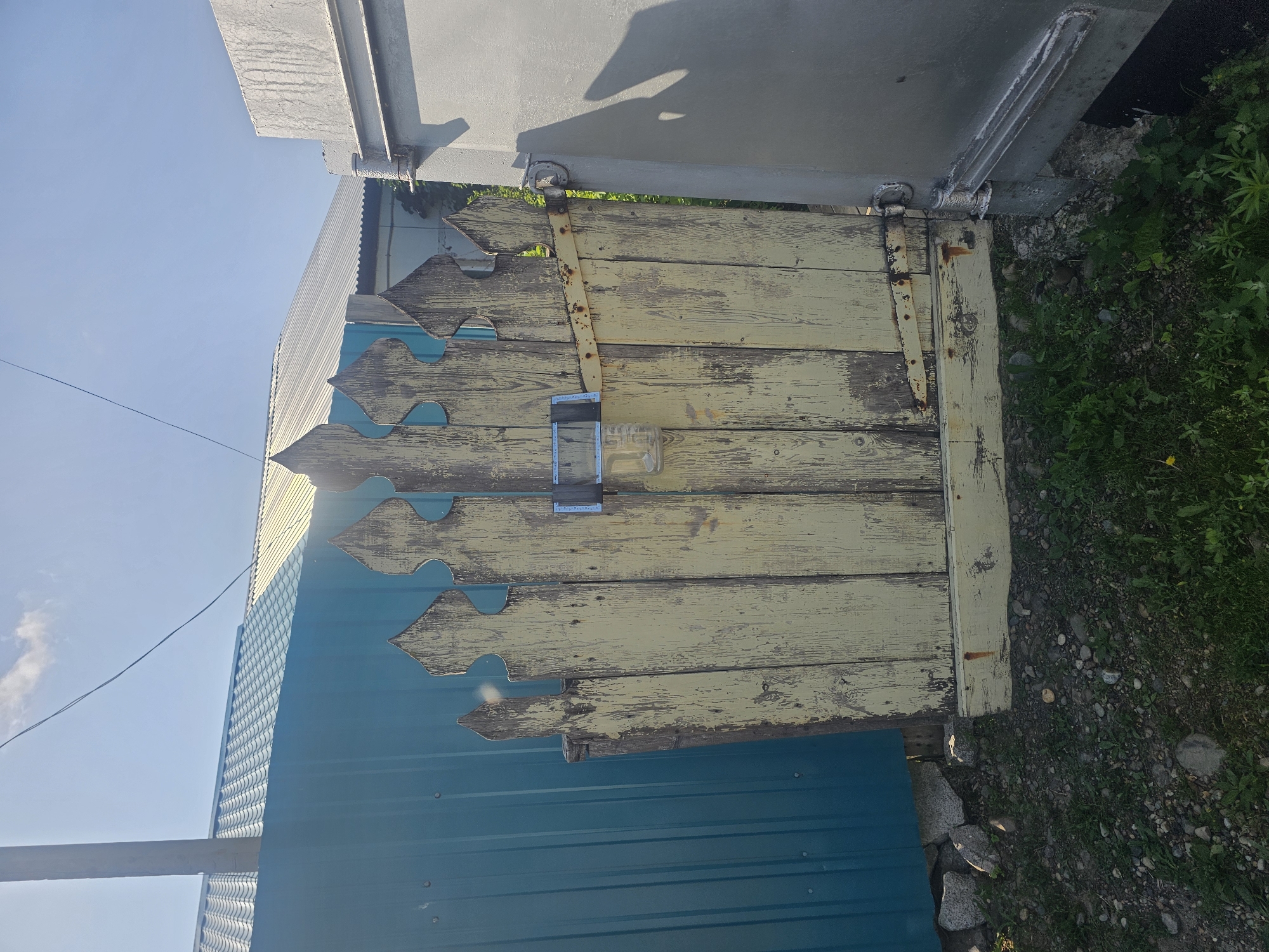 In Georgievka, Russia, an old, weathered yellow wooden gate stands ajar. The gate is adorned with decorative pointed finials along its top edge. Attached to the front of the gate is a clear plastic bottle, secured with tape, suggesting it might be used for collecting something or as a makeshift container. A rusted metal bar crosses the gate horizontally, serving as a decorative element or perhaps reinforcing the structure. The gate is hinged to a grey, industrial-looking structure on the right side of the frame. To the left of the gate, a tall, teal-colored corrugated metal fence stretches into the background. Behind the fence, a house with a silver, corrugated metal roof is partially visible, and a utility pole stands to the left. The ground in front of the gate is covered with a mixture of gravel, rocks, and green grass. The sky above is clear and blue, with a few wispy white clouds. The lighting suggests it is a sunny day.