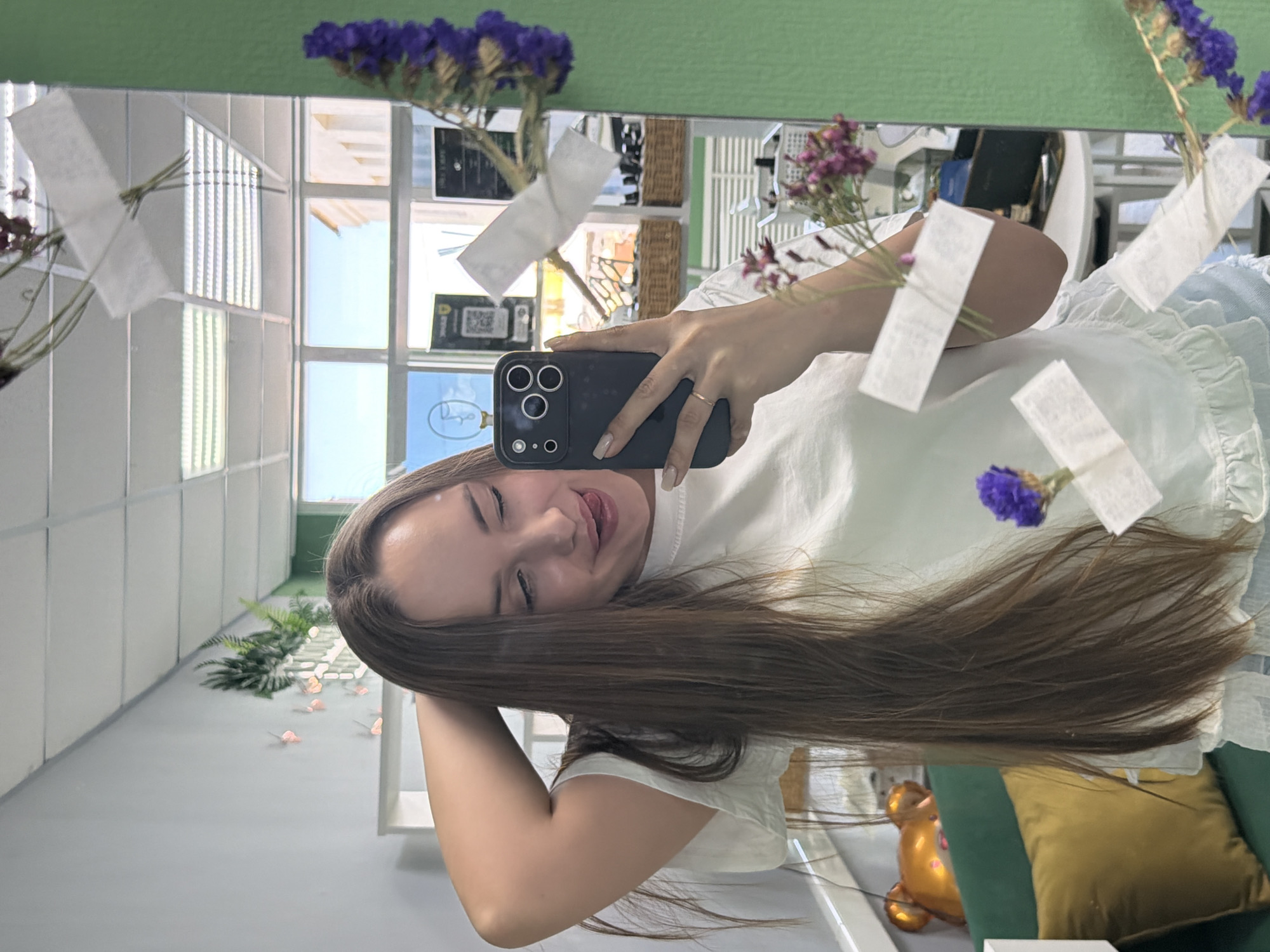 A woman is taking a selfie in front of a mirror in a building in Chaikovsky, Russia. The woman has long brown hair, is wearing a white shirt, and holding an iPhone in her right hand. The mirror reflects a well-lit room with a window and several decorations. The decorations include blue flowers and small white pieces of paper hanging from the ceiling. A green chair with a gold pillow is visible in the background.