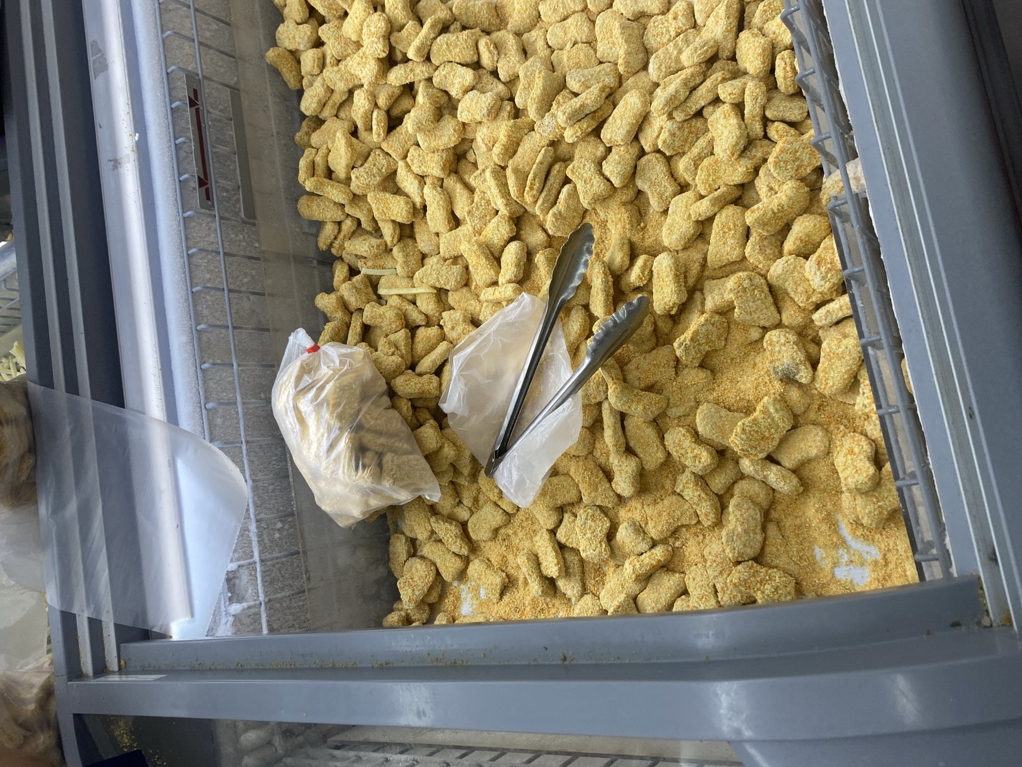 A close-up, high-angle shot shows a large quantity of frozen chicken nuggets in a supermarket freezer. The nuggets are a light golden brown and appear to be breaded. They are piled high in the freezer compartment, with some loose breading scattered on the bottom. Two plastic bags, one open and one sealed, are visible amongst the nuggets, along with a pair of metal tongs resting on the open bag. The freezer itself is a commercial unit with gray metal framing and a wire rack visible at the back. The background is blurred and out of focus, suggesting the photo was taken inside a store. This scene is typical of the frozen food section of a supermarket, which can be found in cities such as Watampone, Indonesia.