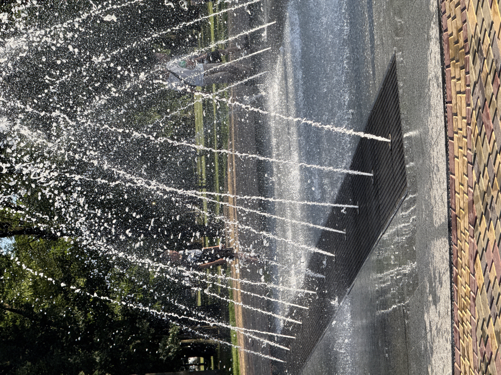 In an outdoor urban park, a series of water jets erupt from a dark grate set into a paved surface. The jets form thin, vertical streams of water that shimmer and refract light, creating a curtain of droplets. Behind the water, two individuals are partially visible. The person on the left, a young male, stands facing forward, wearing a dark t-shirt and shorts, with his face obscured by the spray. To his right, another person, likely an adult male, is walking away from the camera, dressed in a light-colored shirt and pants, carrying a bag over his shoulder. The background reveals lush green trees, suggesting a park setting on a sunny day. The foreground is paved with colorful interlocking bricks.