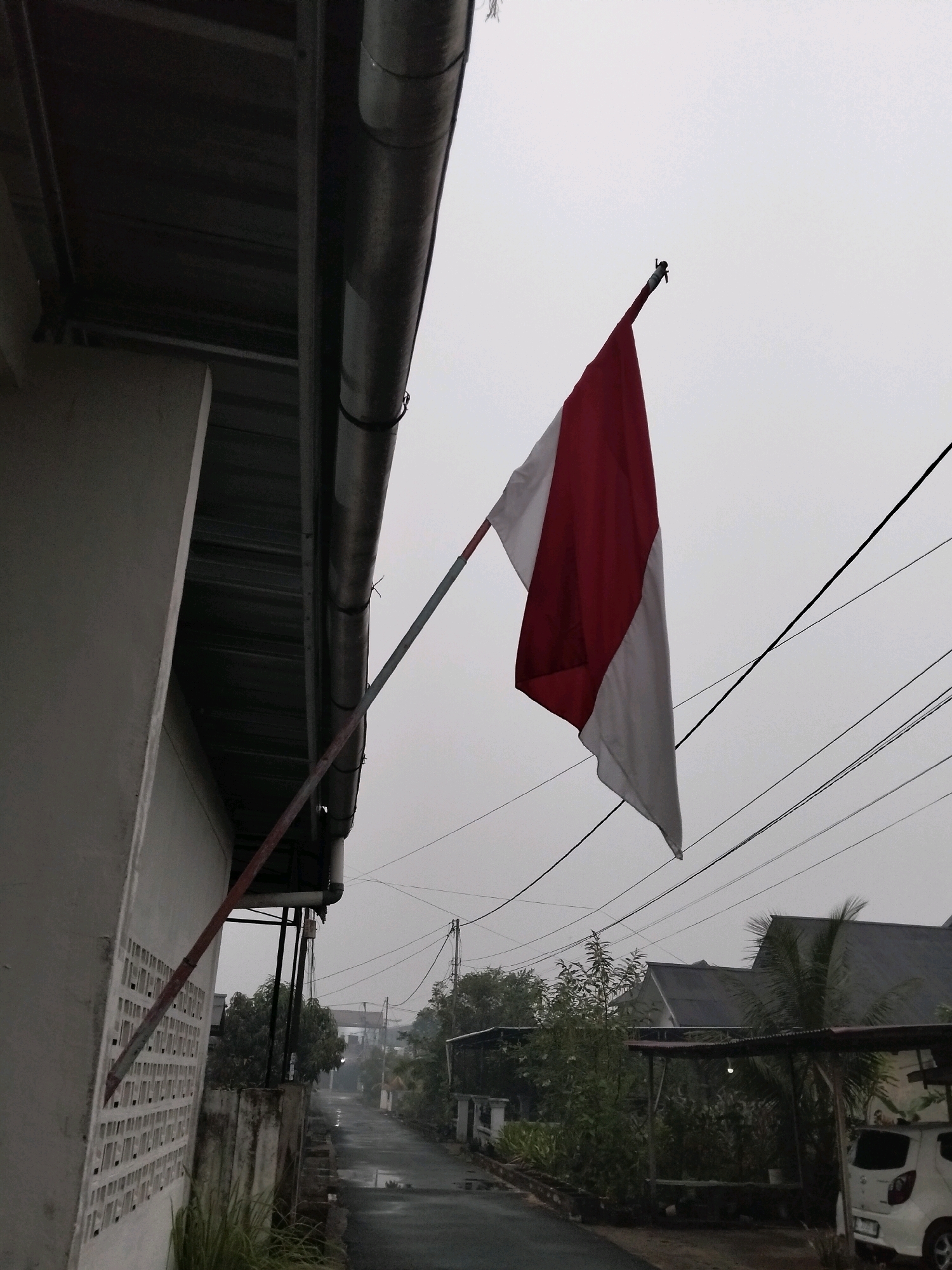 This image depicts an outdoor scene on a cloudy day, likely in Arang Limbung, Indonesia. In the foreground, the side of a white building with a decorative lattice pattern is visible, with a red and white pole extending from it and holding up the Indonesian flag. The flag, with its red upper stripe and white lower stripe, is partially unfurled and hangs against the overcast sky. To the right of the flag, a series of dark electrical wires crisscross the sky. In the background, a narrow, wet asphalt road leads into the distance, flanked by houses with dark roofs and lush green trees. A white car is parked on the right side of the road. The overall atmosphere is subdued, possibly indicating damp weather or early morning fog.