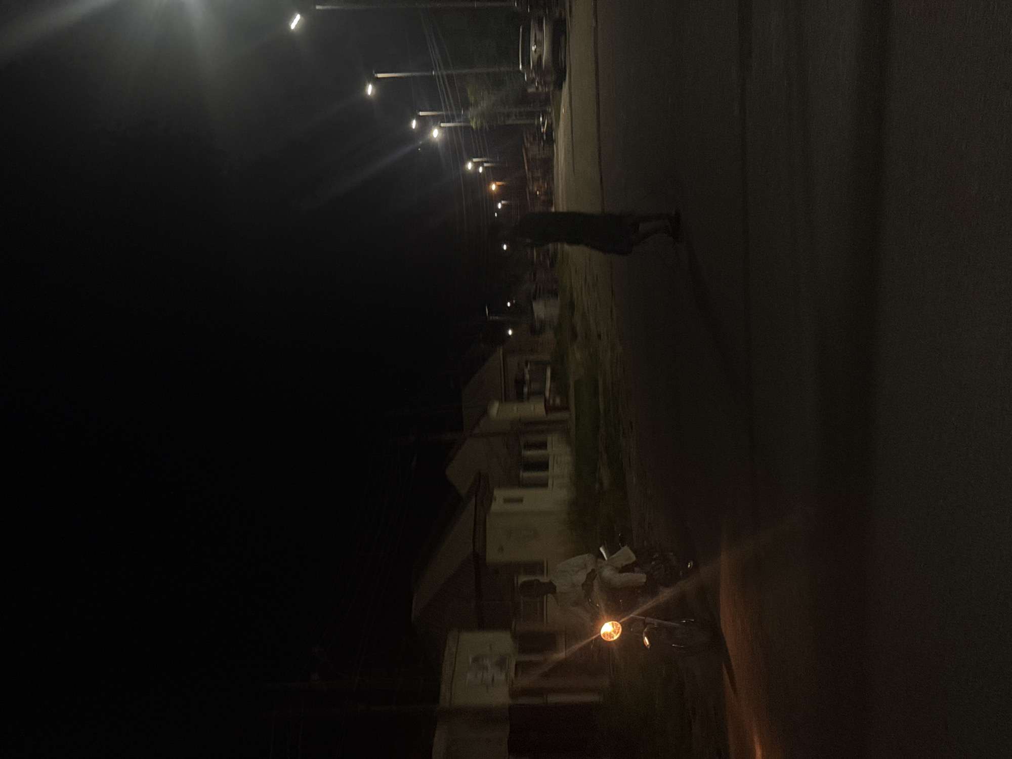 In Abuja, Nigeria, at nighttime, a person rides a motorcycle down a street illuminated by streetlights and the motorcycle's headlight. The rider, dressed in white, is positioned on the left side of the frame, with the motorcycle's headlight casting a bright orange glow. In the background, several single-story buildings with lit windows are visible, suggesting a residential area. Further down the street, another person stands in the road, silhouetted against the distant lights. A car is parked on the right side of the road. The overall scene depicts a quiet, dimly lit urban environment.