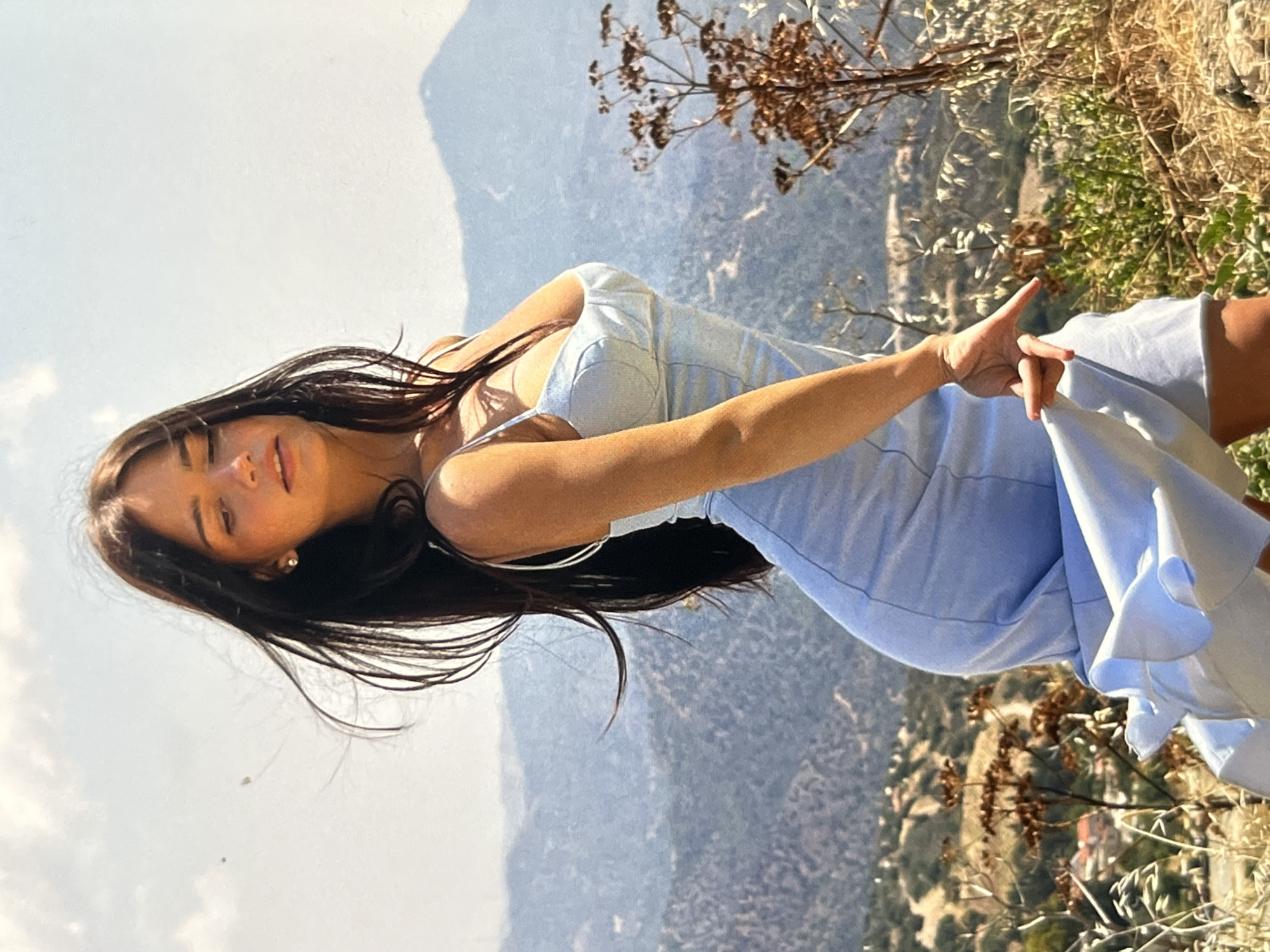 A young woman with long, dark hair is posing outdoors on a hillside. She is wearing a light blue, spaghetti-strap dress. Her body is angled, with her right arm extended and her hand reaching down towards the hem of her dress. Her head is tilted down and to the side, with her eyes closed. The background is a vista of rolling, barren hills and mountains under a bright, partly cloudy sky. Dry brush and some dead plants are in the foreground and scattered throughout the landscape.