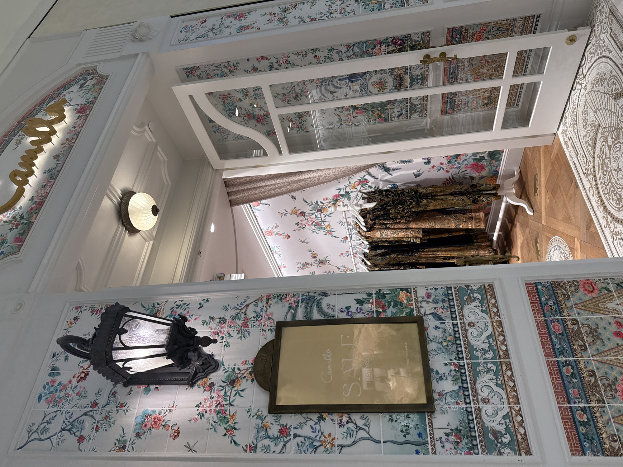 This image presents an upward-angled view of the interior of a luxurious retail store, likely a Camilla boutique, in Miami, USA. The setting is elaborately decorated with walls featuring ornate floral and patterned tiles or wallpaper in various sections.
To the left, an illuminated golden sign spells "Camilla" in a stylized script, set within a decorative arched panel adorned with floral motifs. White decorative molding and an air vent are visible above. A modern-style wall sconce with a ribbed glass shade provides light on the left, while a more traditional black lantern-style fixture hangs on a different patterned wall section to the lower left.
In the upper-middle of the frame, white-framed double doors with multiple glass panes are open, revealing more patterned surfaces beyond, including hanging clothes or textiles with intricate designs. The floor visible through the doors appears to be hardwood with a circular patterned inlay.
A prominent gold-framed sign on one of the patterned walls clearly advertises "SALE" in large letters, accompanied by smaller text that reads "IN STORE" and "TERMS & CONDITIONS APPLY." The entire space exudes a high-end, richly decorated aesthetic, characterized by a profusion of colorful patterns and detailed architectural elements. No individuals are present in the scene. The lighting suggests an interior during operating hours, without external weather indications.