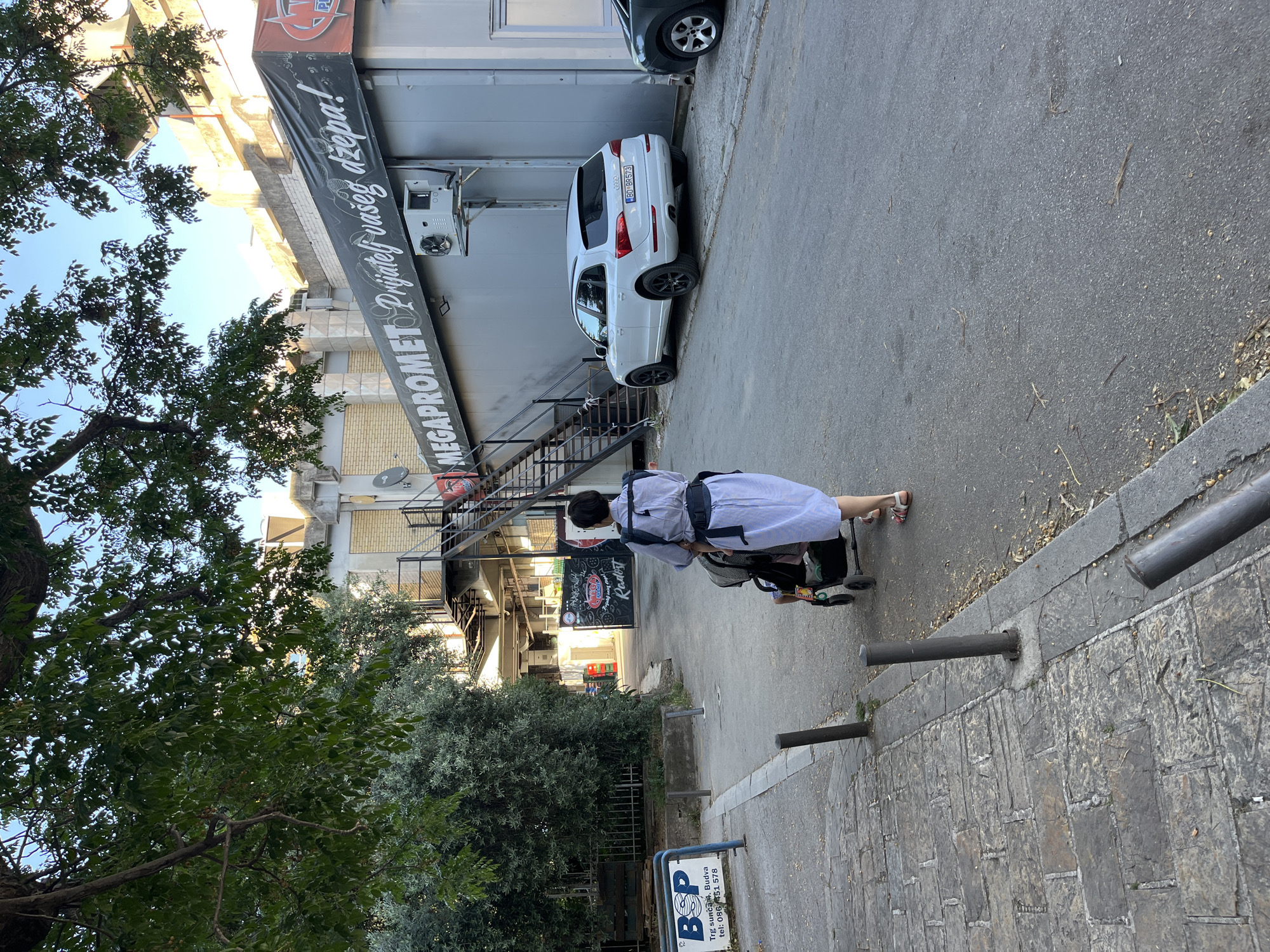 A medium shot captures a woman, seen from the back, pushing a stroller along a sidewalk. She is dressed in a light blue striped shirt and pink pants. The sidewalk is made of grey rectangular paving stones. Metal posts act as barriers along the sidewalk. A sign reading "BOP Trg sunca 4. Budva tel: 086 451 578" is visible on the left. The backdrop features a building with a large banner that reads "MEGAPROMET Prijatelj vaseg džepa!" A white Audi SUV with license plate BD BR573 is parked next to the building. The surroundings include trees. A sign reading "Radost" is also visible in the background. The setting appears to be in Budva, Montenegro, given the location details on the sign.
