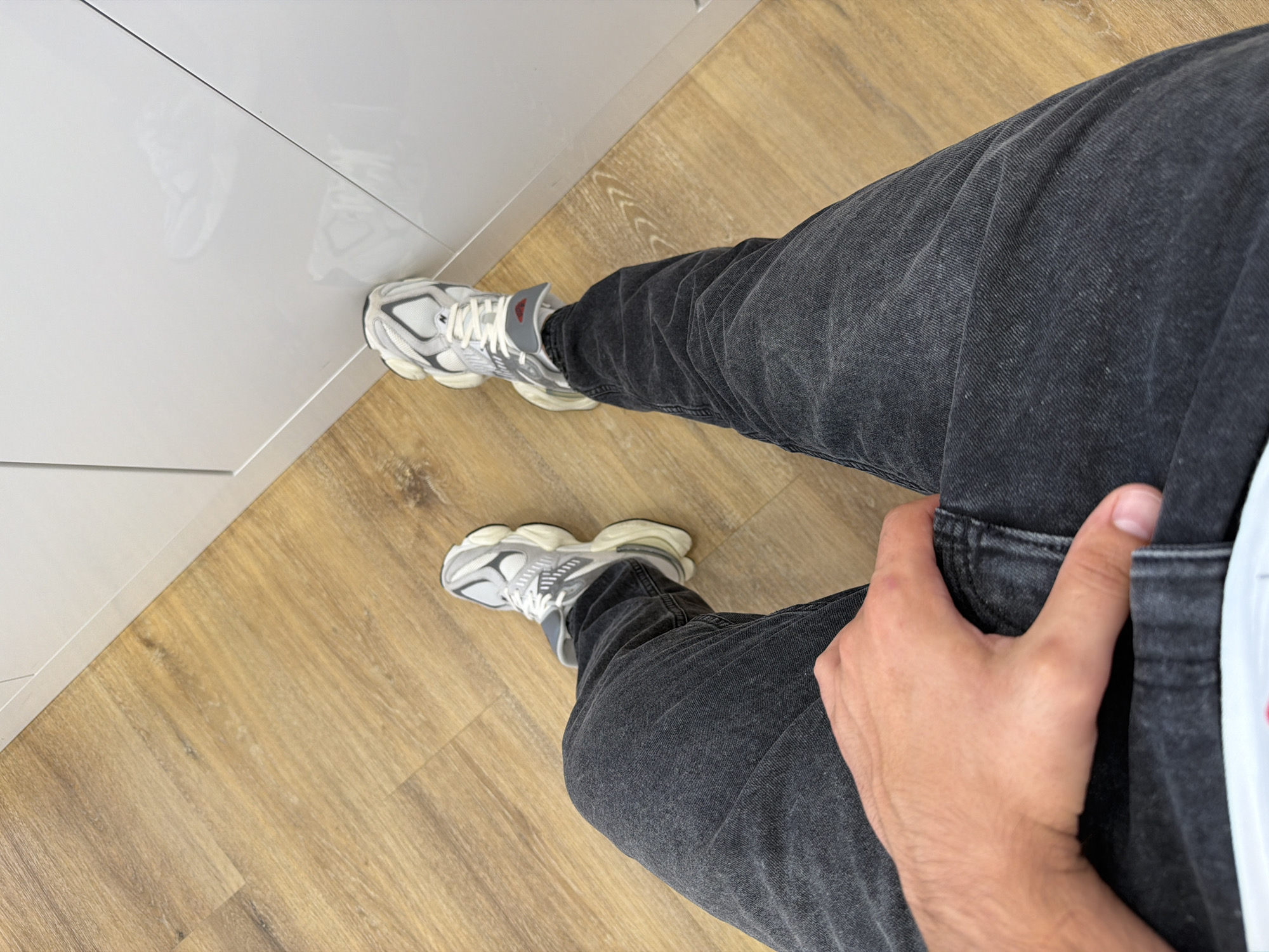The image presents a top-down view of a person's lower body, focused on their legs and feet, in an indoor setting. The individual wears dark grey or faded black denim jeans, which appear slightly creased, and a pair of light grey and white athletic sneakers. The sneakers feature a distinctive chunky sole and mesh detailing, with a visible "NB" logo on the side, indicating they are New Balance shoes. A small red patch, possibly with text, is present on the tongue of the shoe but is illegible.
One hand, with light skin tone, is seen resting on the upper thigh of the right leg. The flooring is a light-colored wood-effect laminate or similar material, and a clean, glossy white surface, likely cabinetry or furniture, runs along the left side of the frame, showing faint reflections of the shoes. The lighting is bright and even, typical of indoor illumination, and does not suggest a specific time of day. The scene depicts a static pose, with no discernible activity or event taking place. The overall impression is a modern, minimalist indoor environment, located in Sofia, Bulgaria, as per the provided context. No other explicit text or emotional cues are visible.