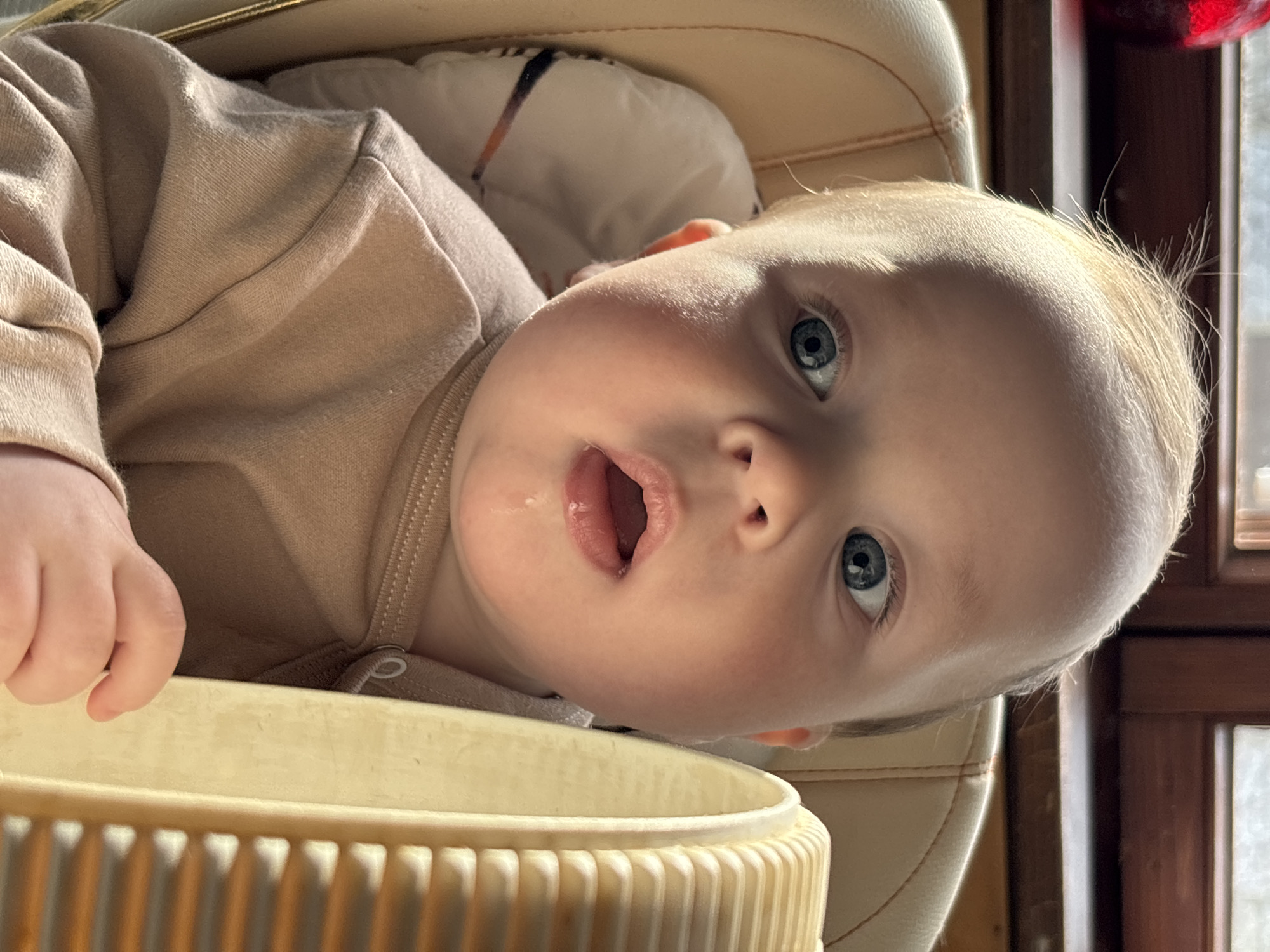 A close-up, slightly tilted image shows a baby seated in a high chair, looking directly at the camera with wide, curious blue eyes. The baby has fair skin and light blonde, wispy hair, catching the bright sunlight which also casts shadows on the face and creates reflections in the eyes. A drop of liquid, likely saliva, is visible on the baby's lower lip, and the mouth is open in an expression of surprise or engagement.
The baby is wearing a plain, long-sleeved top in a warm beige or light brown color. The high chair is a pale cream or yellow color with a ribbed texture on its tray and side. A soft, white pillow or cushion supports the baby's head. The baby's left hand is visible, resting on the high chair tray.
In the background, a window with a dark wooden frame is partially visible, indicating an indoor setting. A small, red, spherical object can be seen near the window in the upper right corner of the frame, possibly a decoration. The strong, natural illumination suggests it is daytime and sunny. No text is visible in the image. The provided location context is Markovskiy, Russia.